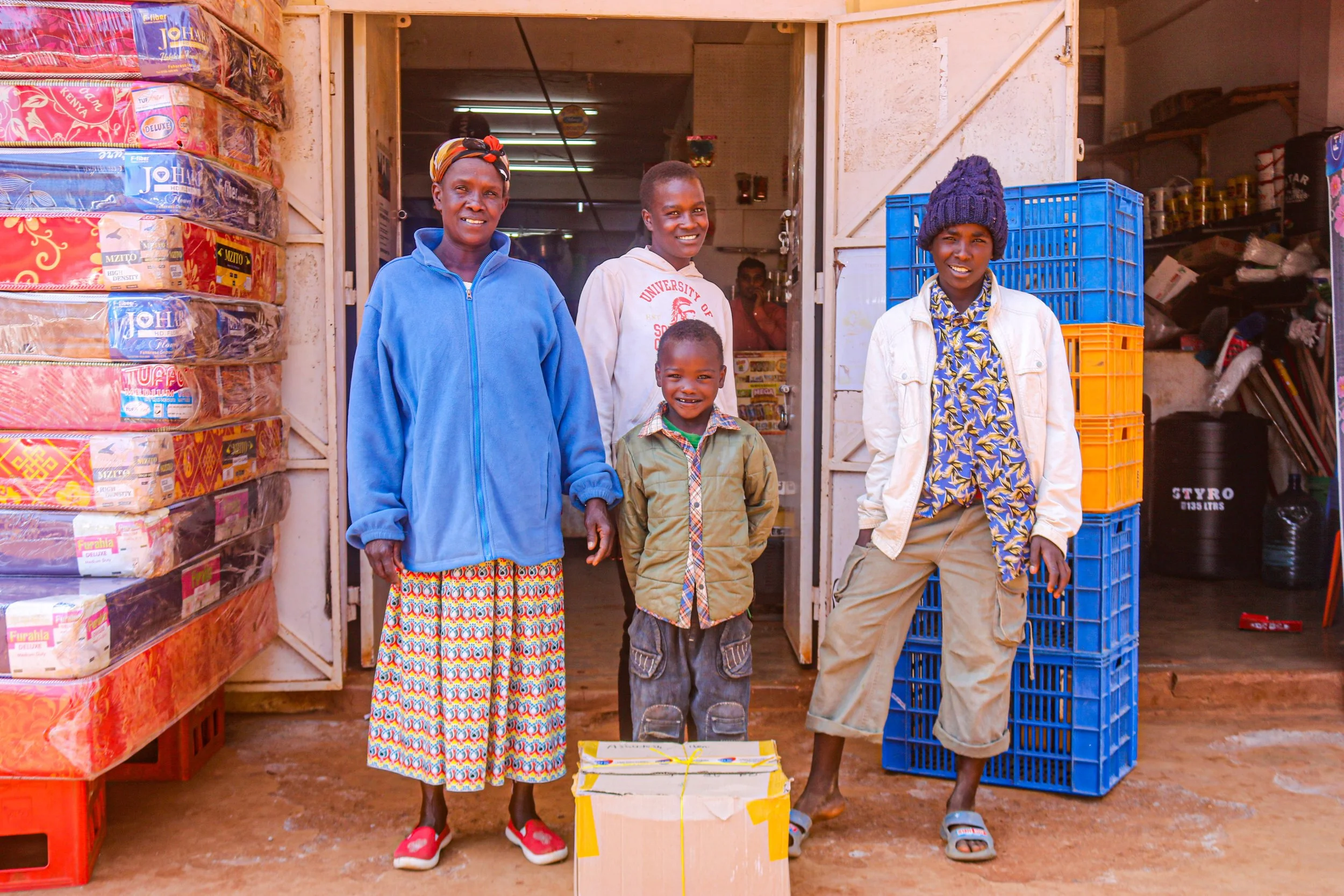 Four people standing outside a small shop, smiling at the camera. The shop has colorful crates and packaged goods visible inside.
