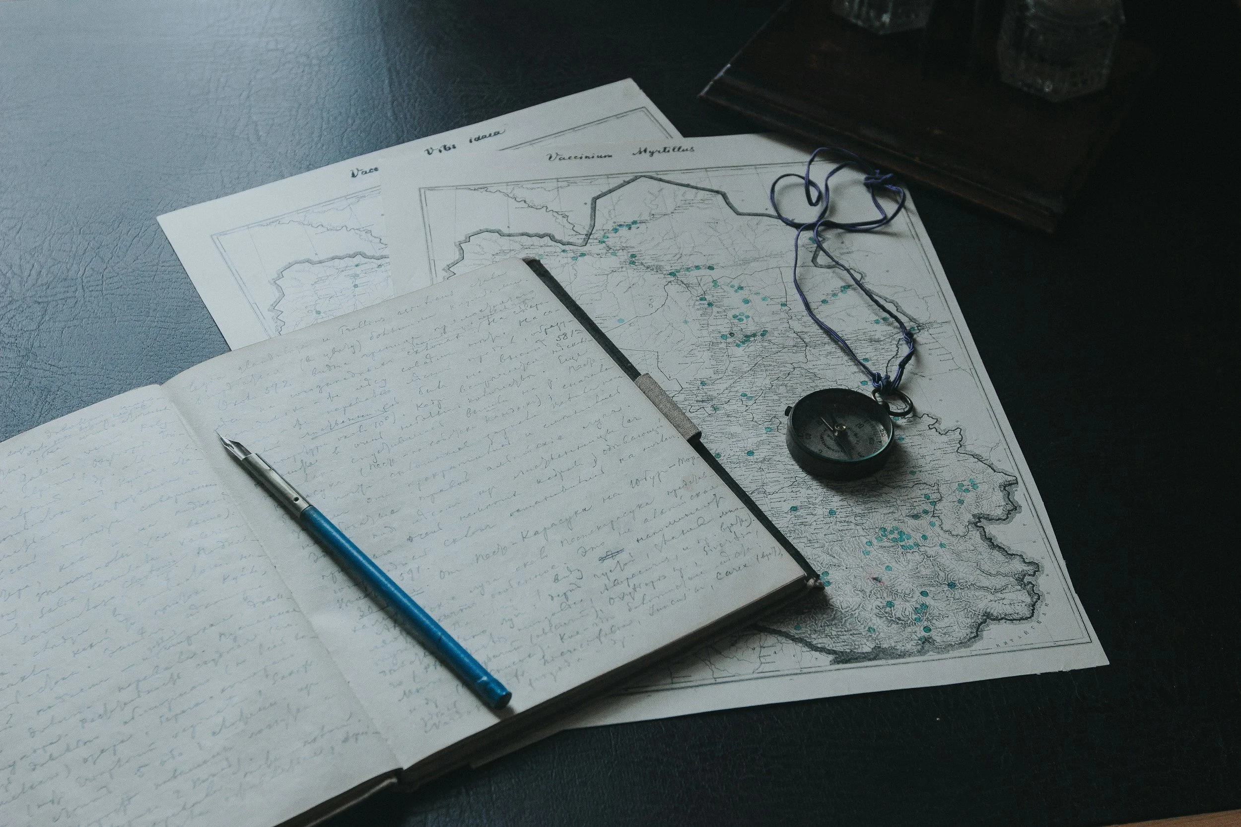 A desk with a handwritten journal, a pen, a map with markers, a compass, and a string with a compass attached.