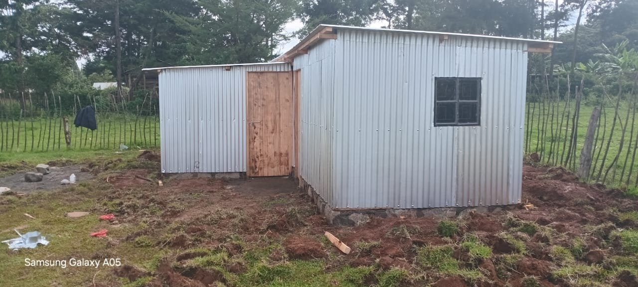 A small corrugated metal building with a wooden door and a window, set on uneven ground in a rural area with trees and a fence in the background.