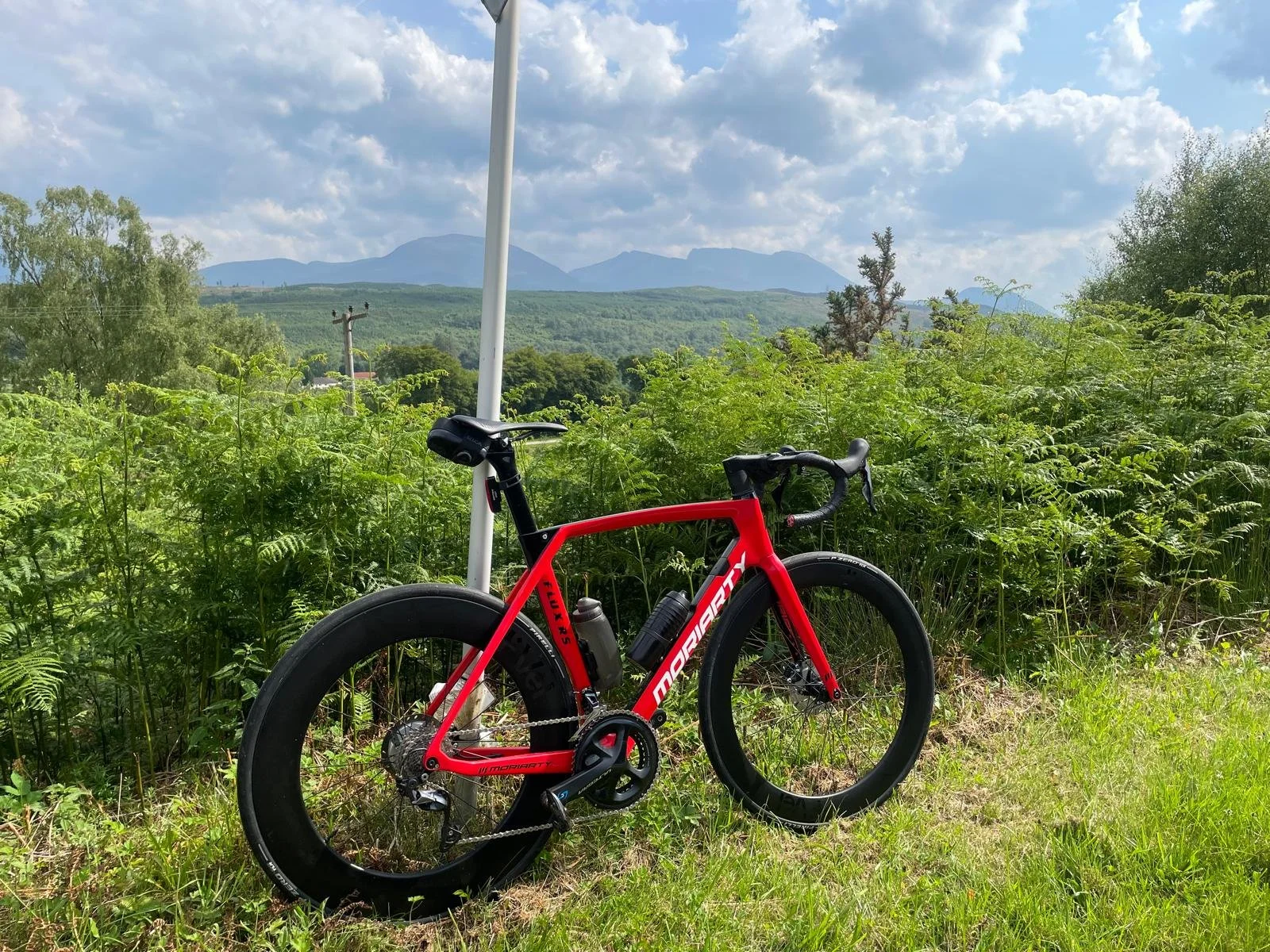 A red and black mountain bike resting against a white pole on a grassy area, with green shrubs and trees, rolling hills, and mountains in the distance under a partly cloudy sky.