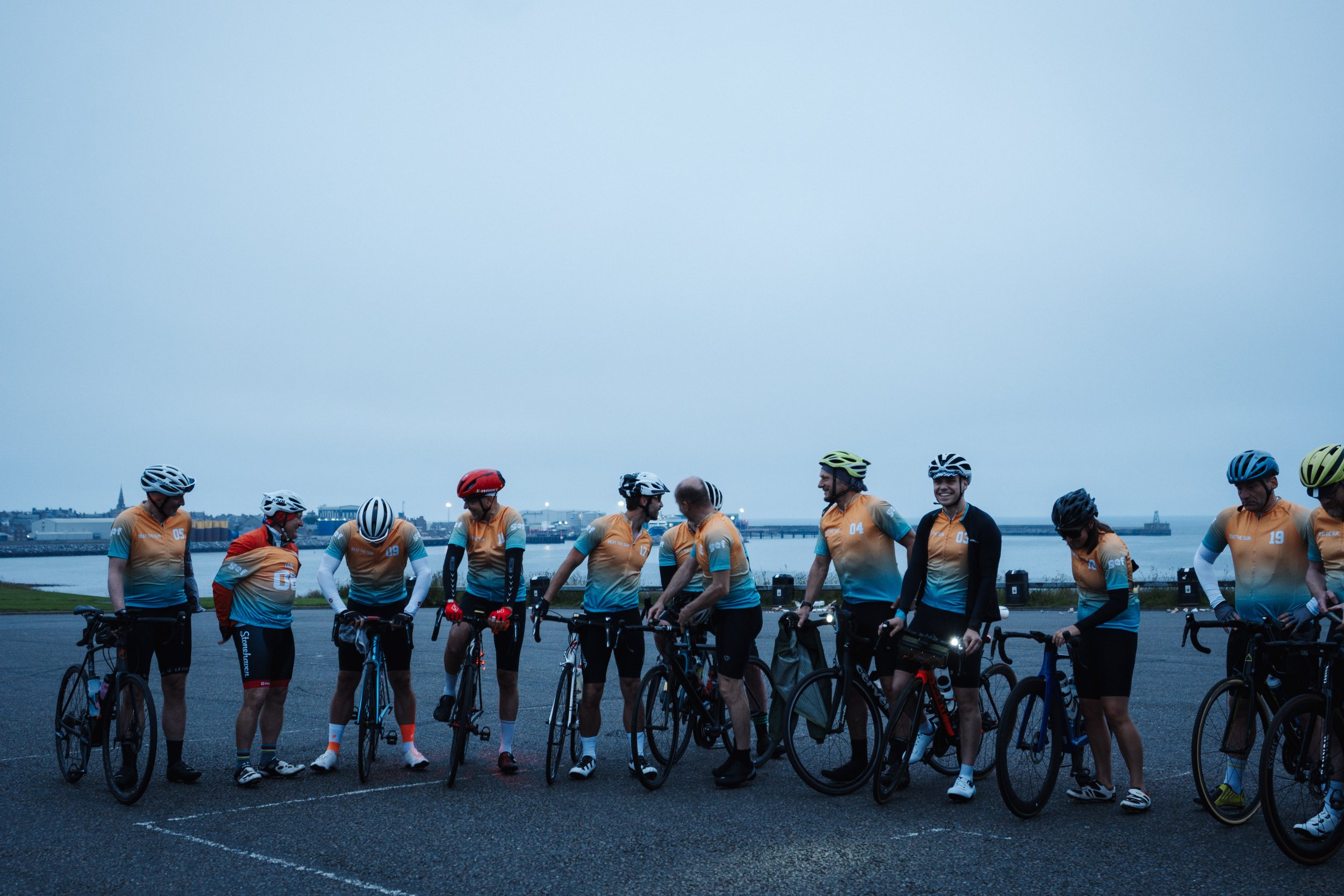 A group of cyclists standing with bikes near a body of water at dusk, wearing matching orange and blue cycling jerseys and helmets.