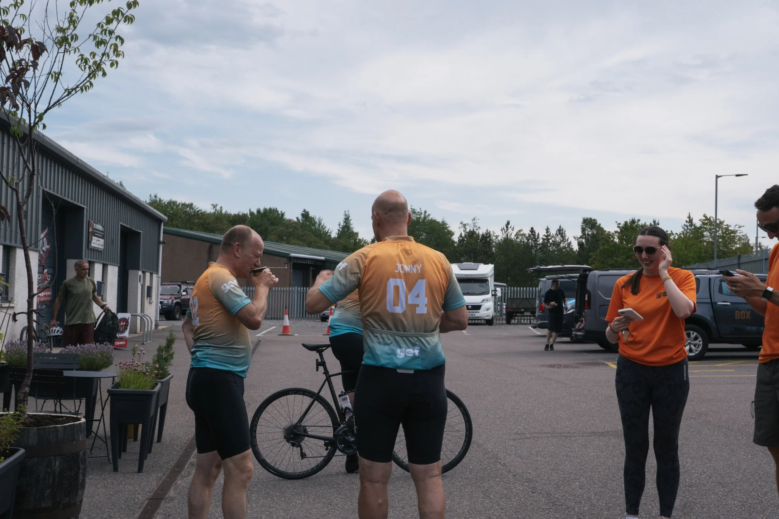 Group of four people in a parking lot, three wearing matching orange and teal cycling jerseys, one woman holding a smartphone, and two men with bicycles.