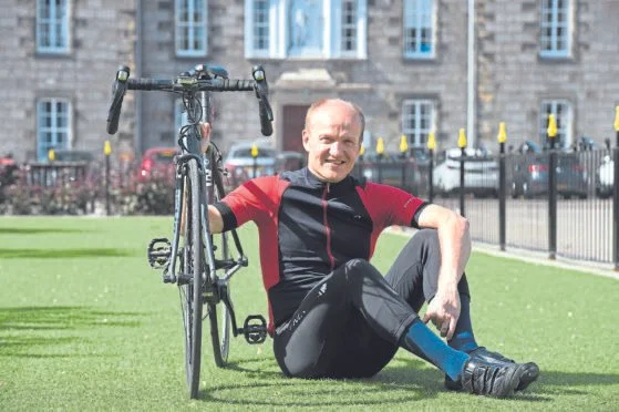 An older man sitting on the grass next to his bicycle, smiling at the camera in a park or outdoor area with a building in the background.