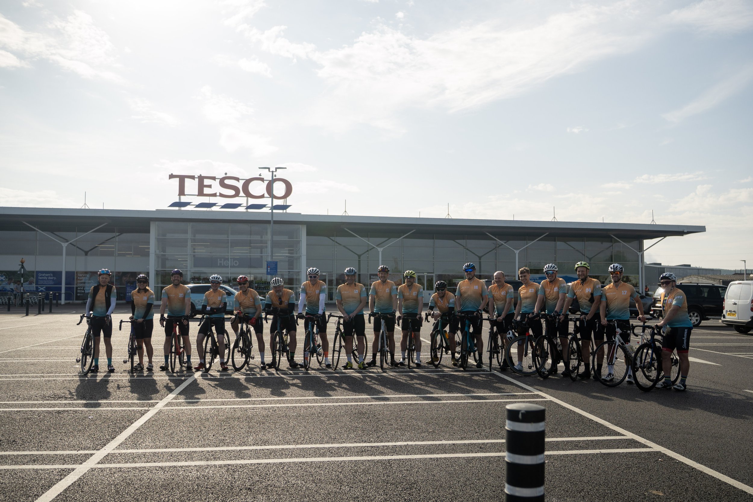 A group of cyclists wearing yellow and blue jerseys stands with their bikes in the parking lot of a Tesco store, with the store's large sign visible in the background.