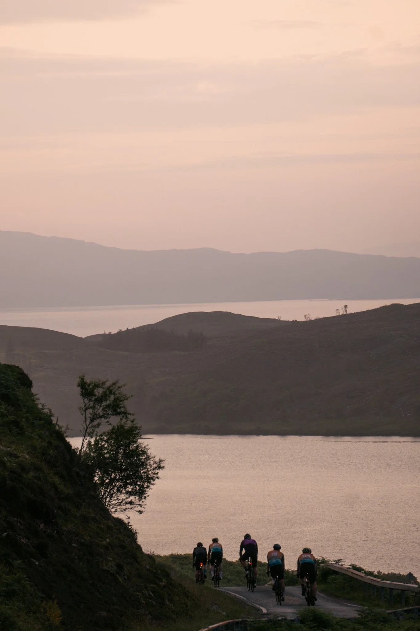 Five cyclists riding along a winding road near a large body of water during sunset, with hills and mountains in the background.
