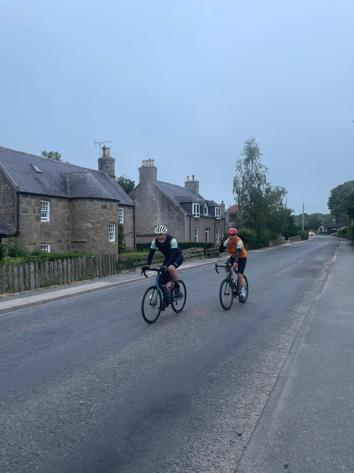 Two cyclists riding on the street in a quiet residential area with stone houses and trees.