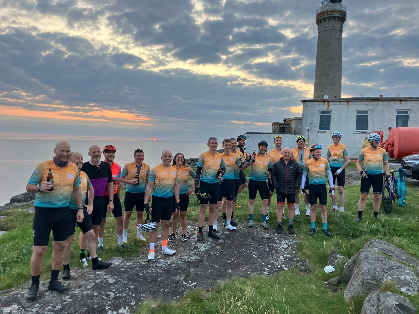 A group of cyclists wearing matching orange and blue jerseys posing together on a grassy area near a lighthouse at sunset.