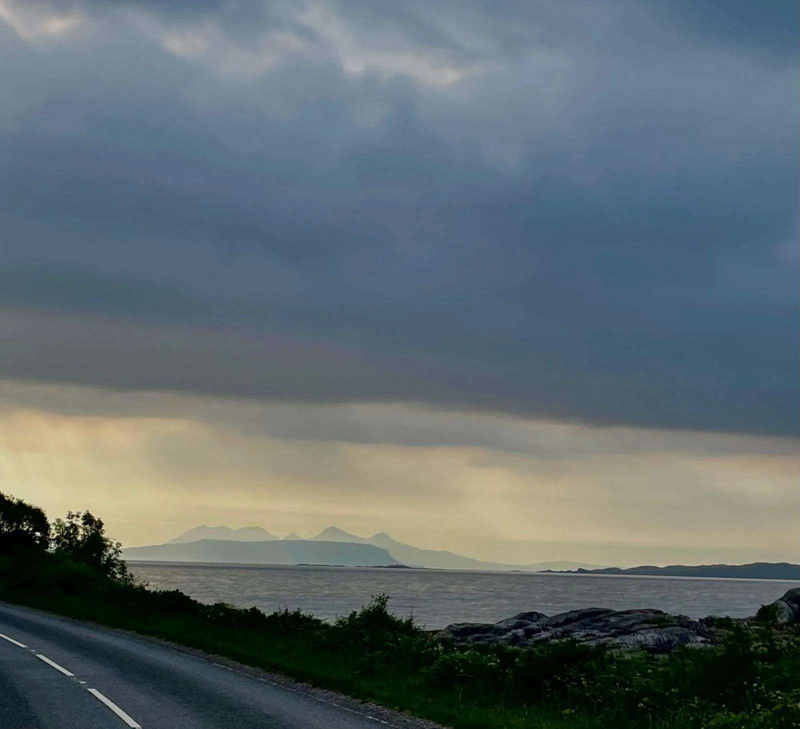 A scenic view of a coastline with mountains in the distance under a cloudy sky, with a road and greenery in the foreground.