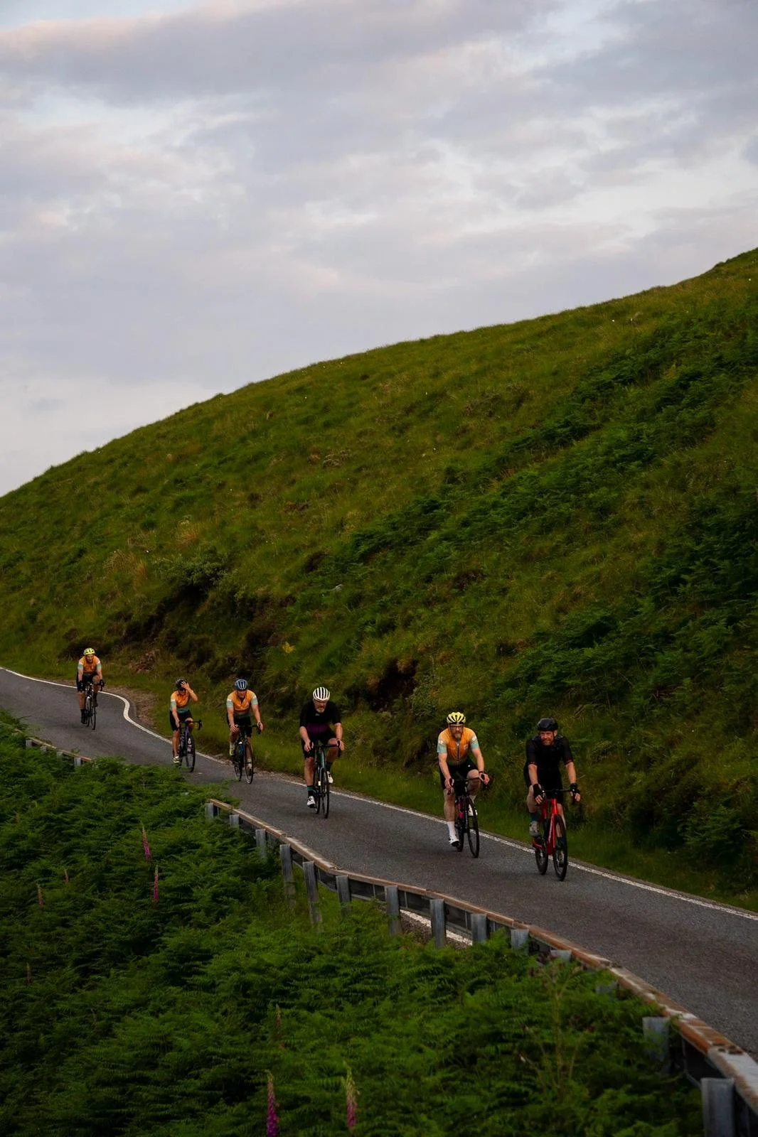 Six cyclists riding up a winding road through green hilly landscape under a cloudy sky.