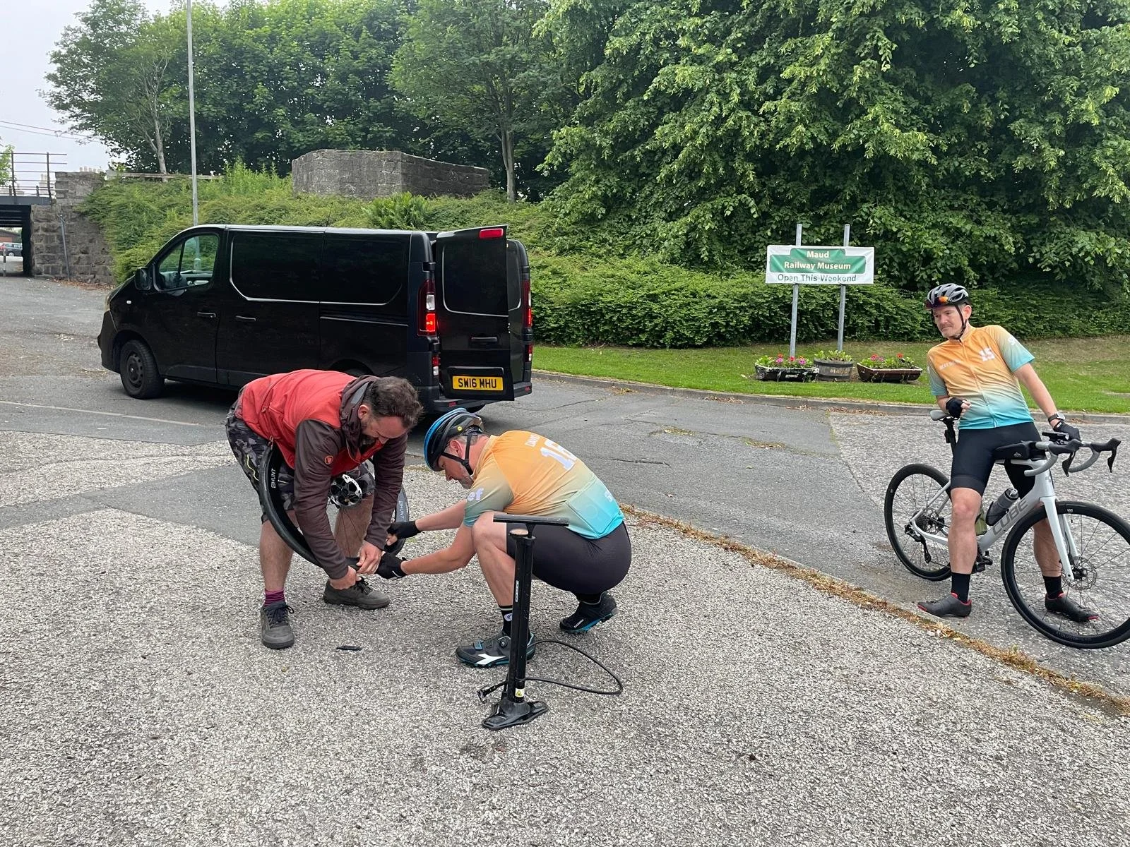 Two men in cycling gear assisting another man in fixing a flat tire on his bicycle on a paved surface, with a black van, green trees, and a sign for the Maud Railway Museum in the background.