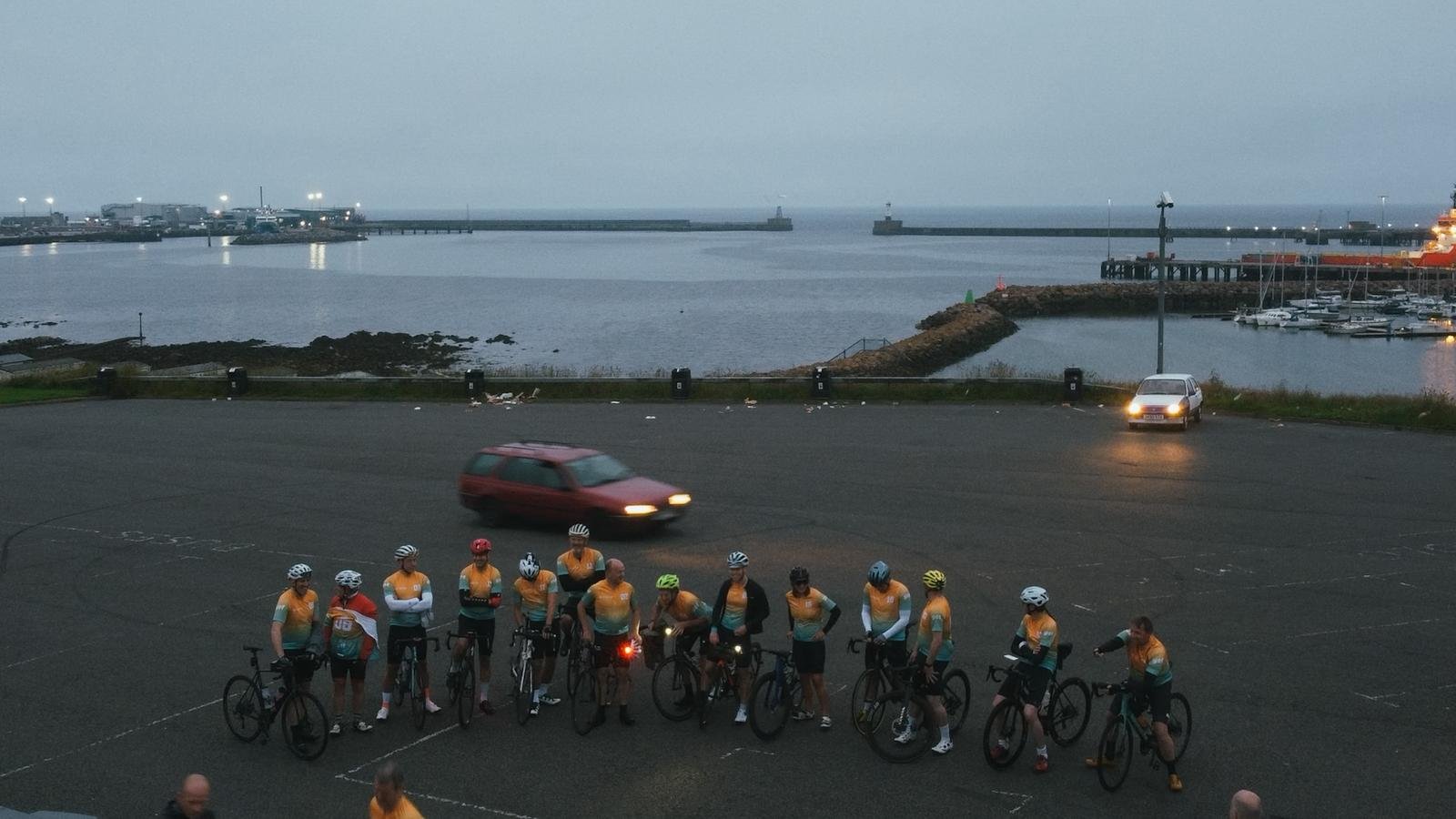 A group of cyclists in orange jerseys and helmets gathered in a parking lot near a harbor with boats and a breakwater, during dusk.