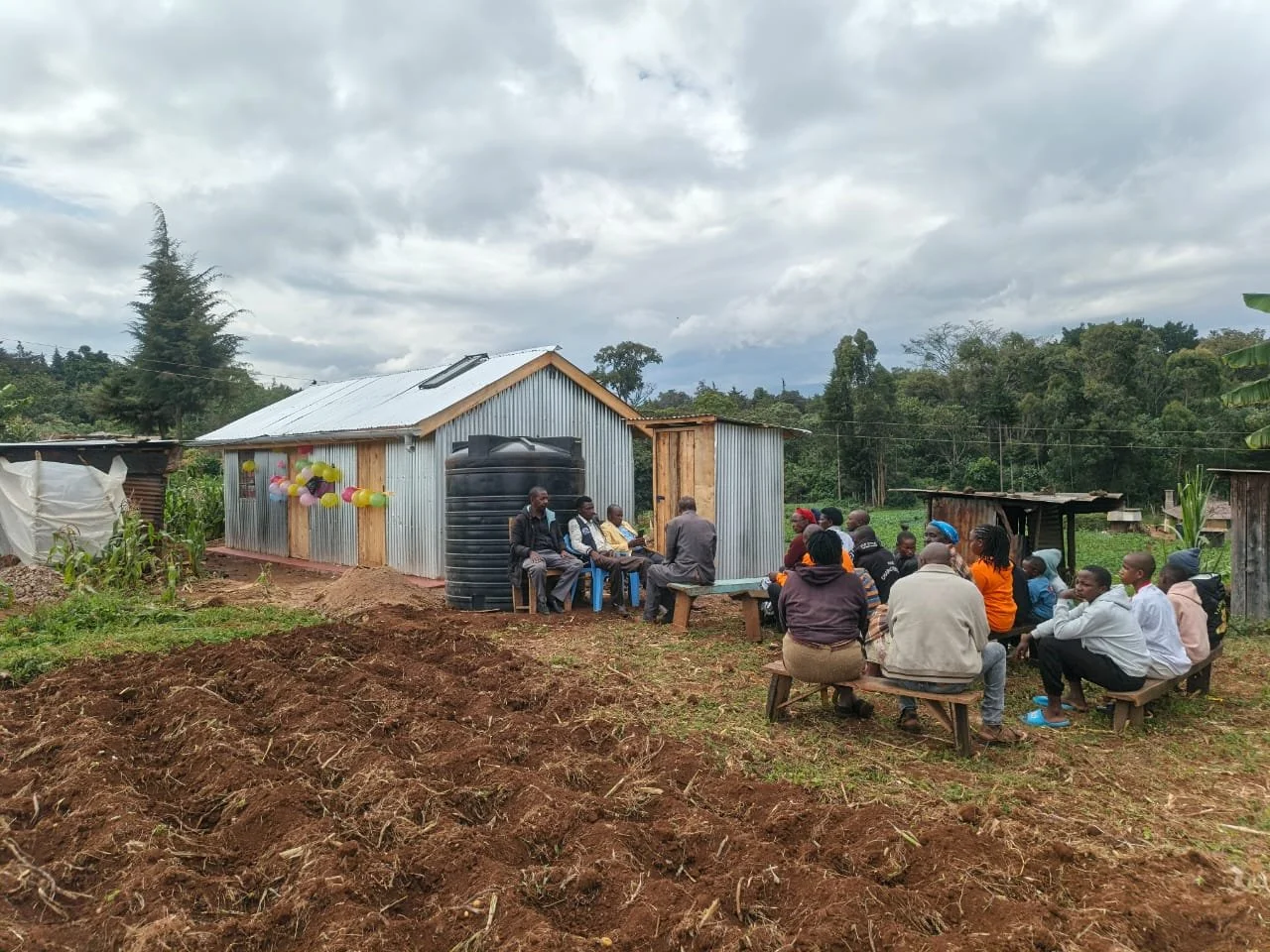 A group of people gathered outside a metal house for a meeting or event, with a cloudy sky overhead.