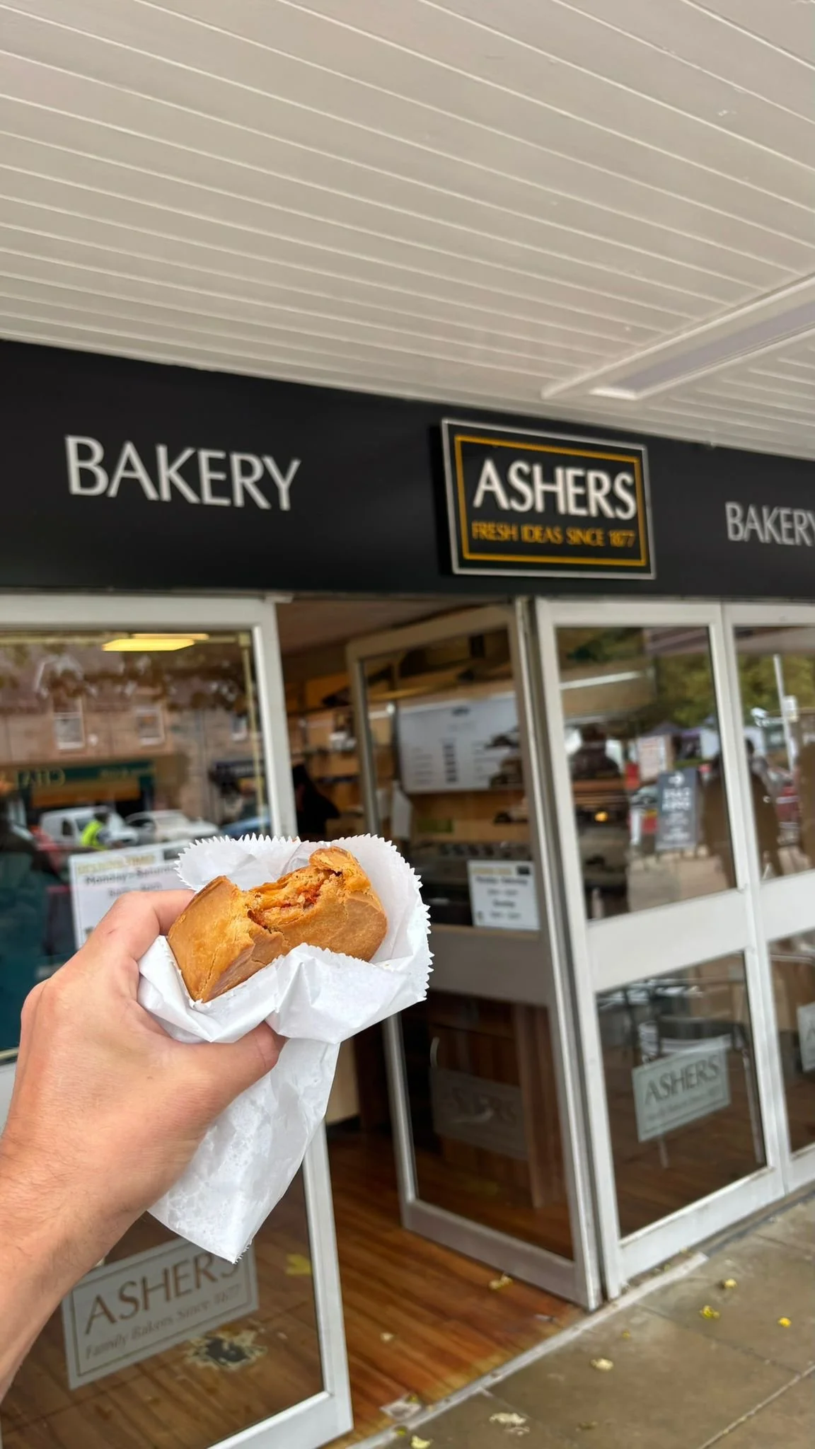 A person holding a partially eaten apple pie in front of Ashers bakery storefront.