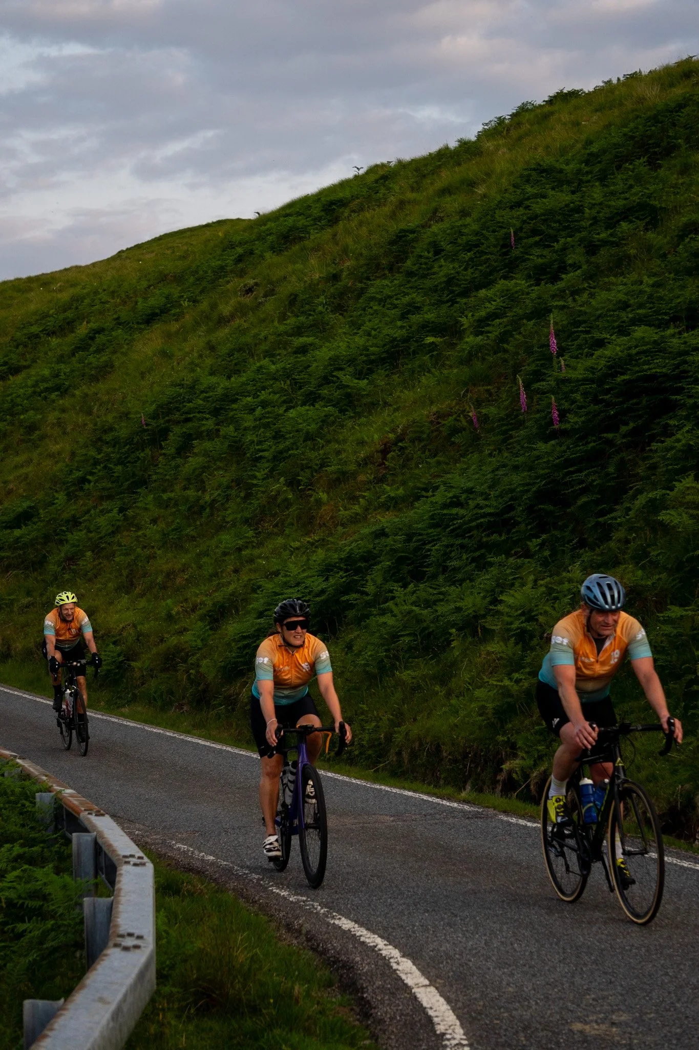 Three cyclists in orange and turquoise jerseys riding uphill on a winding paved road next to a grassy hillside with purple flowers, under a cloudy sky.