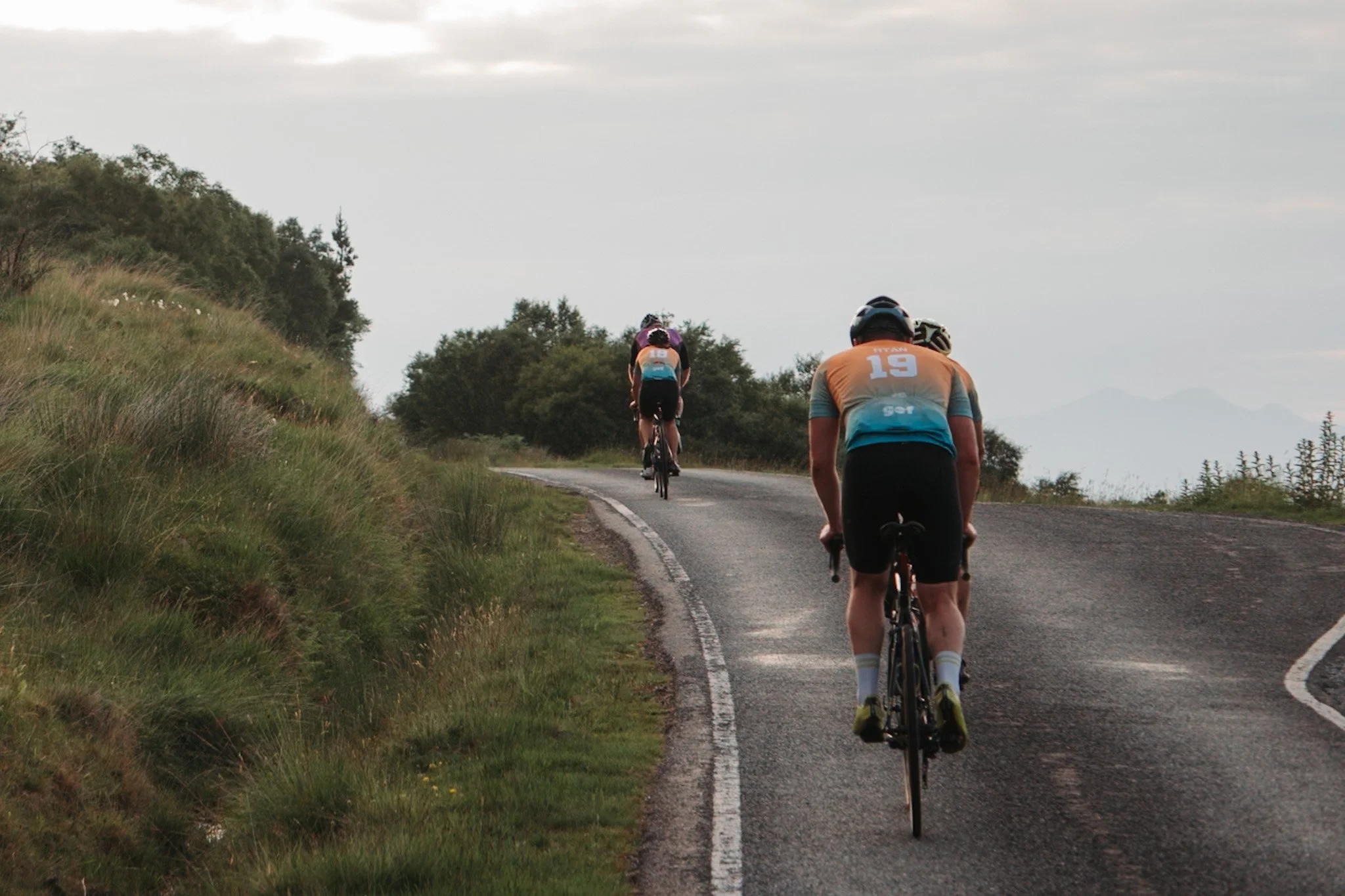 Three cyclists riding uphill on a rural road bordered by grass and bushes.