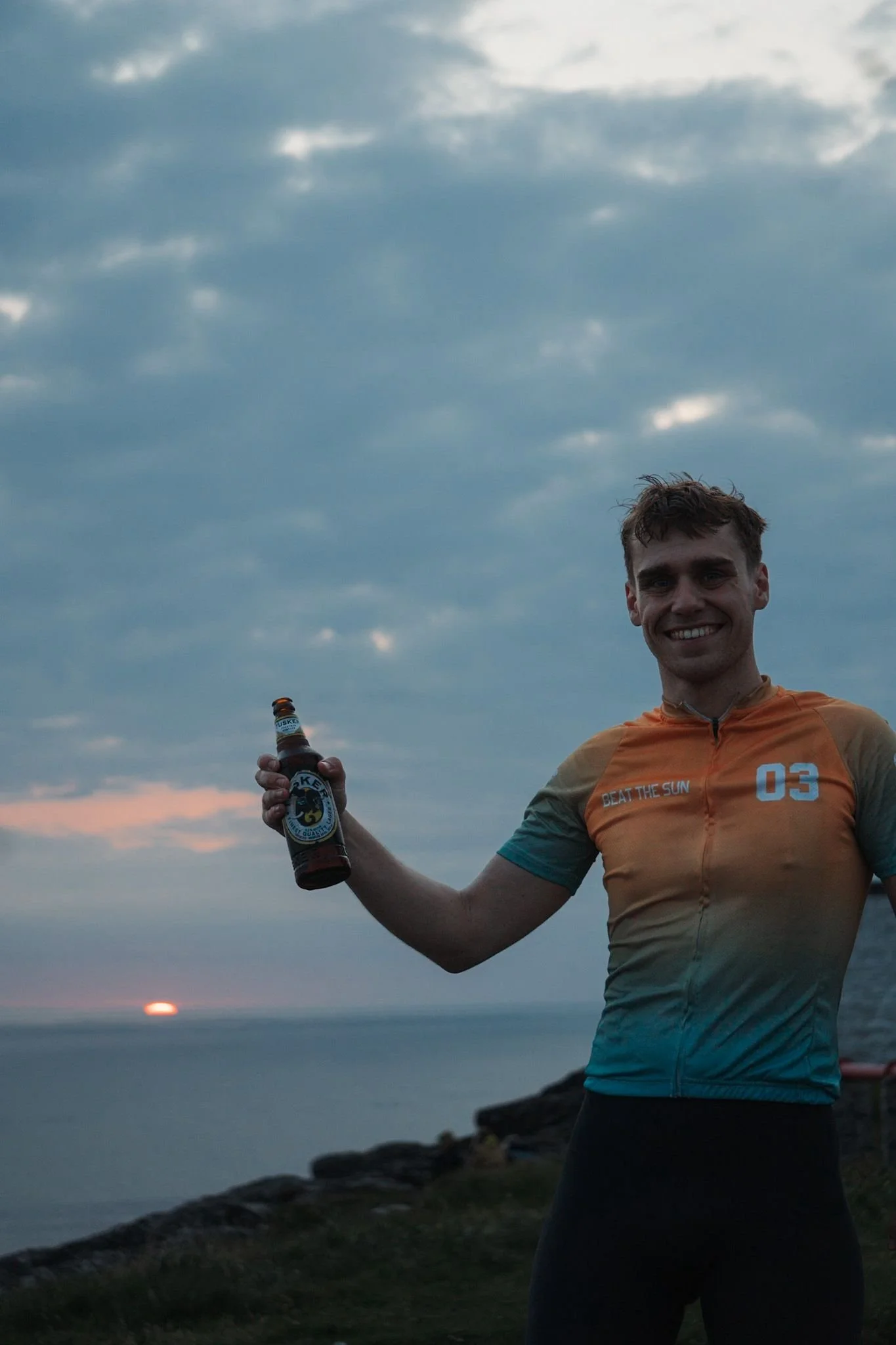 A smiling young man in a colorful cycling jersey holding a beer bottle, standing outdoors near the coast at sunset with a cloudy sky above.
