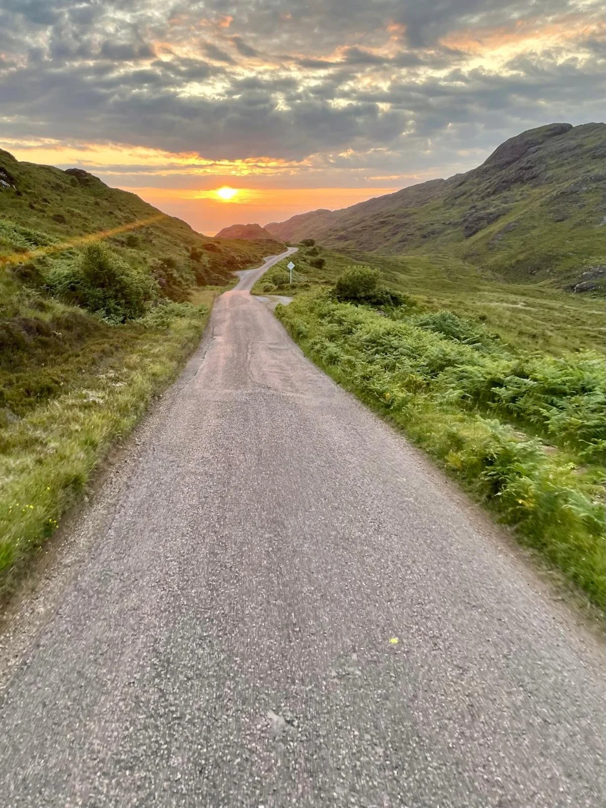 A winding gravel road through green hills at sunset with clouds in the sky.