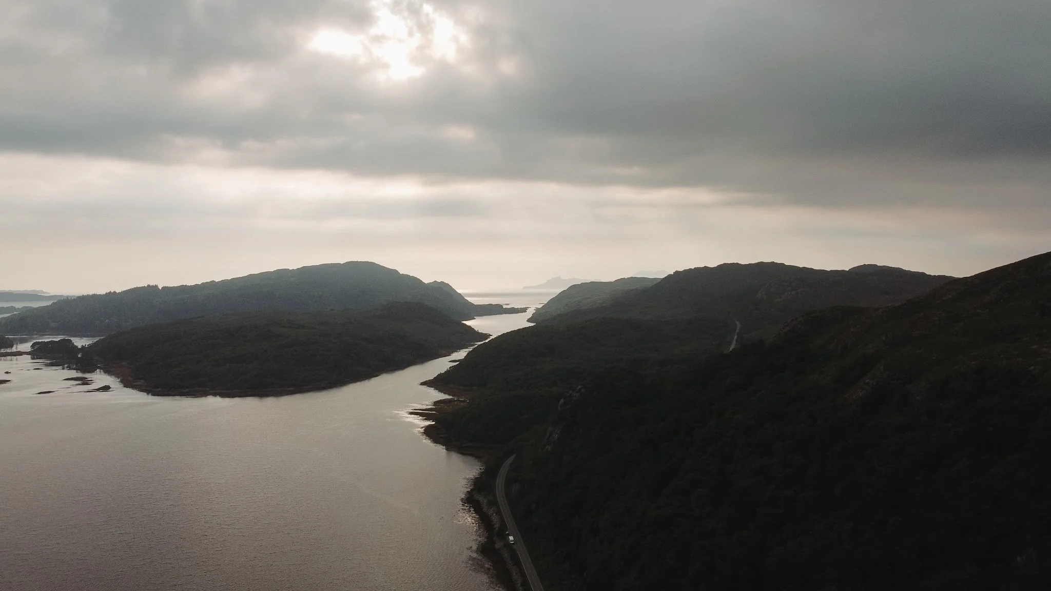 A landscape view of a winding river flanked by dark, hilly terrain under a cloudy sky.