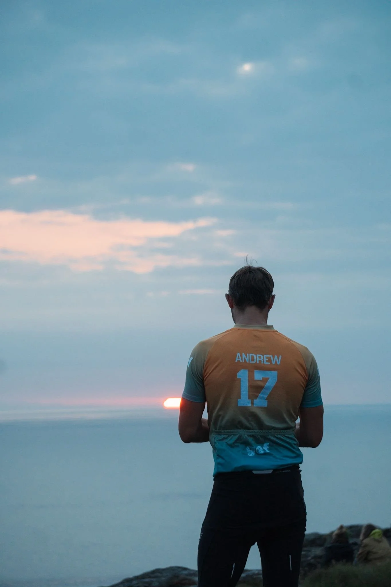 Man in sports jersey with the name Andrew and number 17 on the back, standing on rocky terrain near water, watching the sunset.