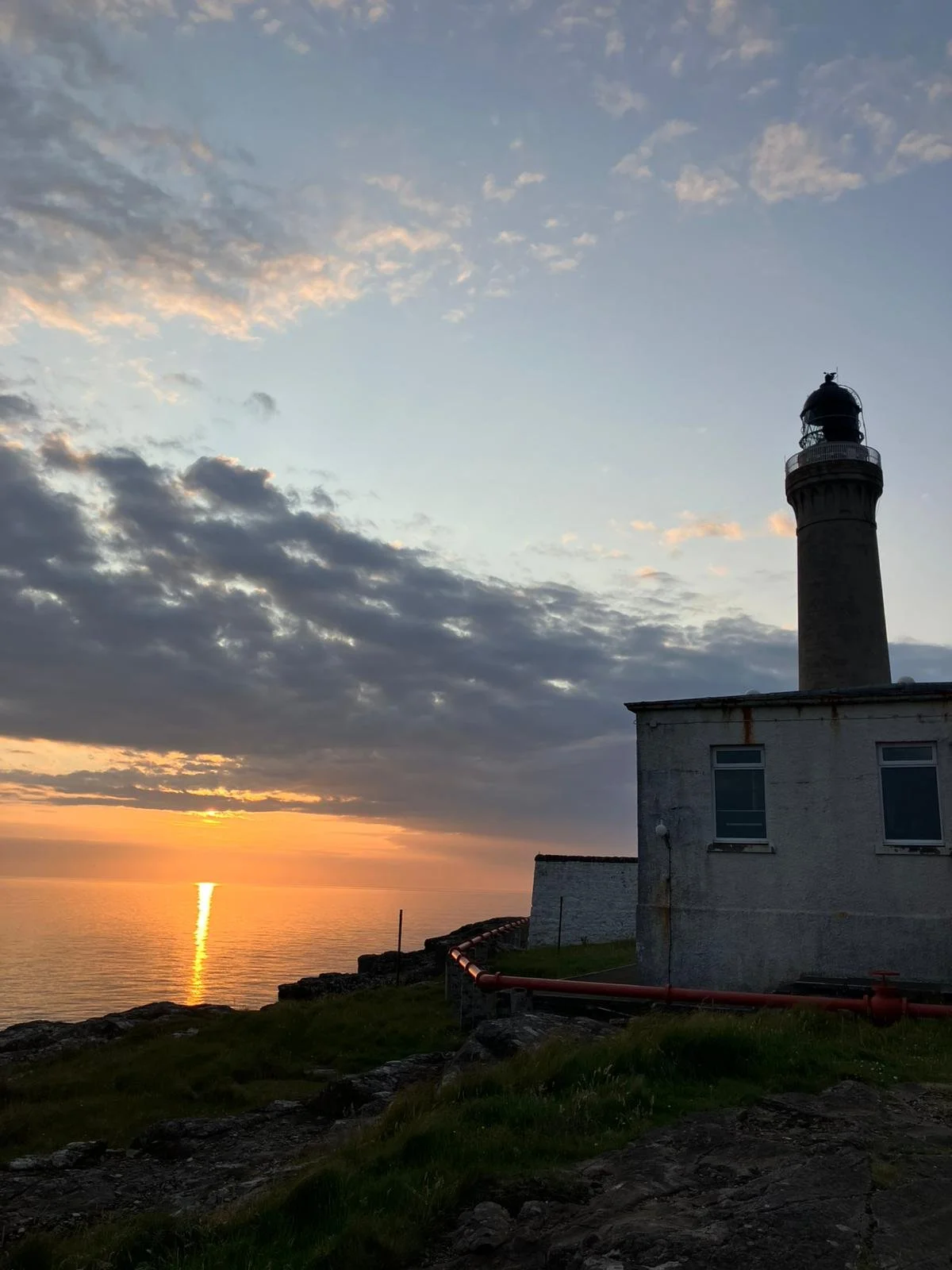 A lighthouse standing on a rocky coastline with a sunset over the ocean in the background.