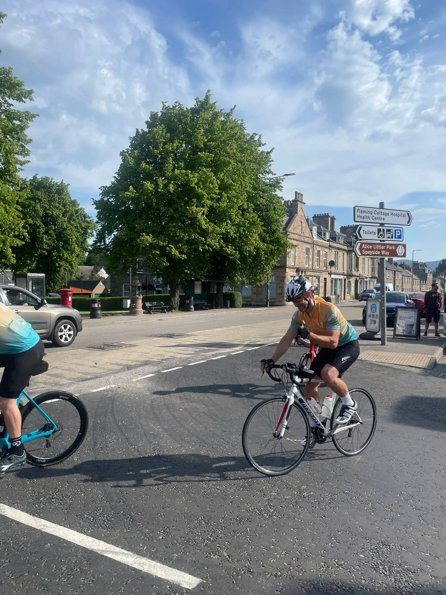 Cyclists riding on a city street with trees, historic buildings, and directional signs in the background.