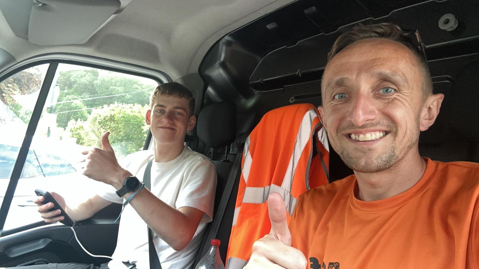 Two men sitting inside a vehicle, both smiling and giving thumbs-up gestures. The man in the foreground is wearing an orange shirt and the man in the background is holding a phone and wearing a white shirt.