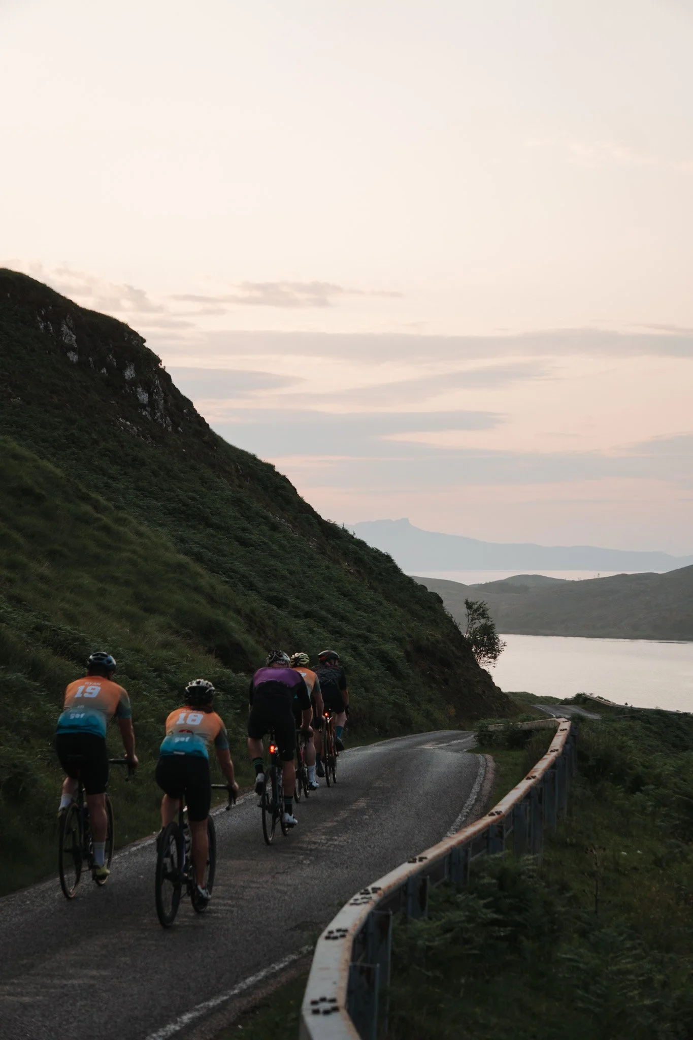 A group of six cyclists riding on a winding road along a hillside during dusk, with water and mountains in the distance.