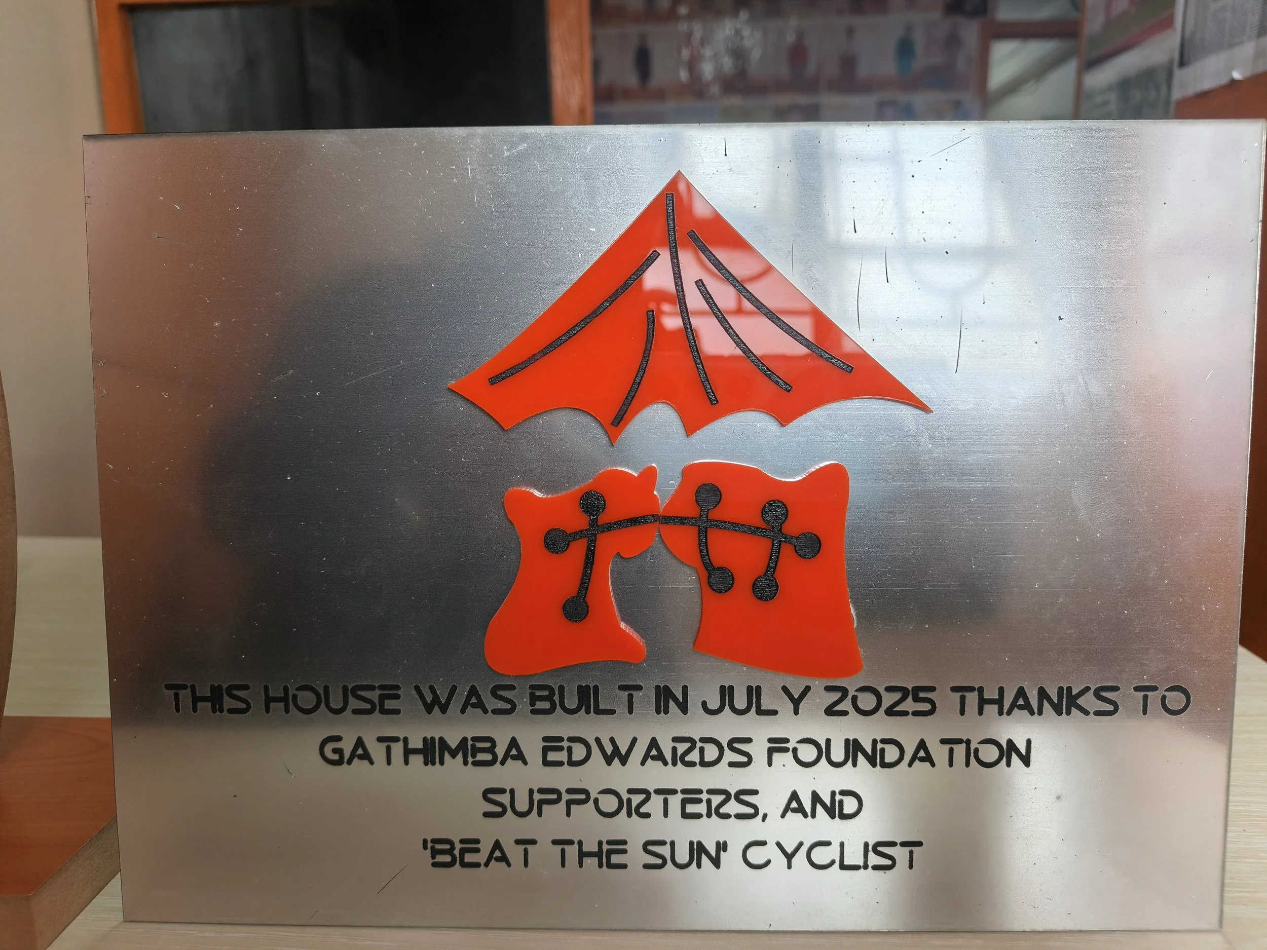 A metal plaque with an orange graphic of a house and a cyclist, and text that reads: "This house was built in July 2025 thanks to Gathimba Edwards Foundation supporters, and 'Beat the Sun' cyclist."