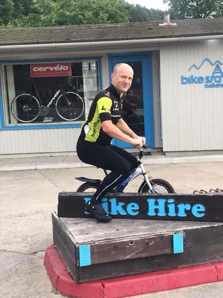 A man in cycling attire sitting on a small bicycle on an elevated wooden platform labeled 'Bike Hire' outside a bike shop.