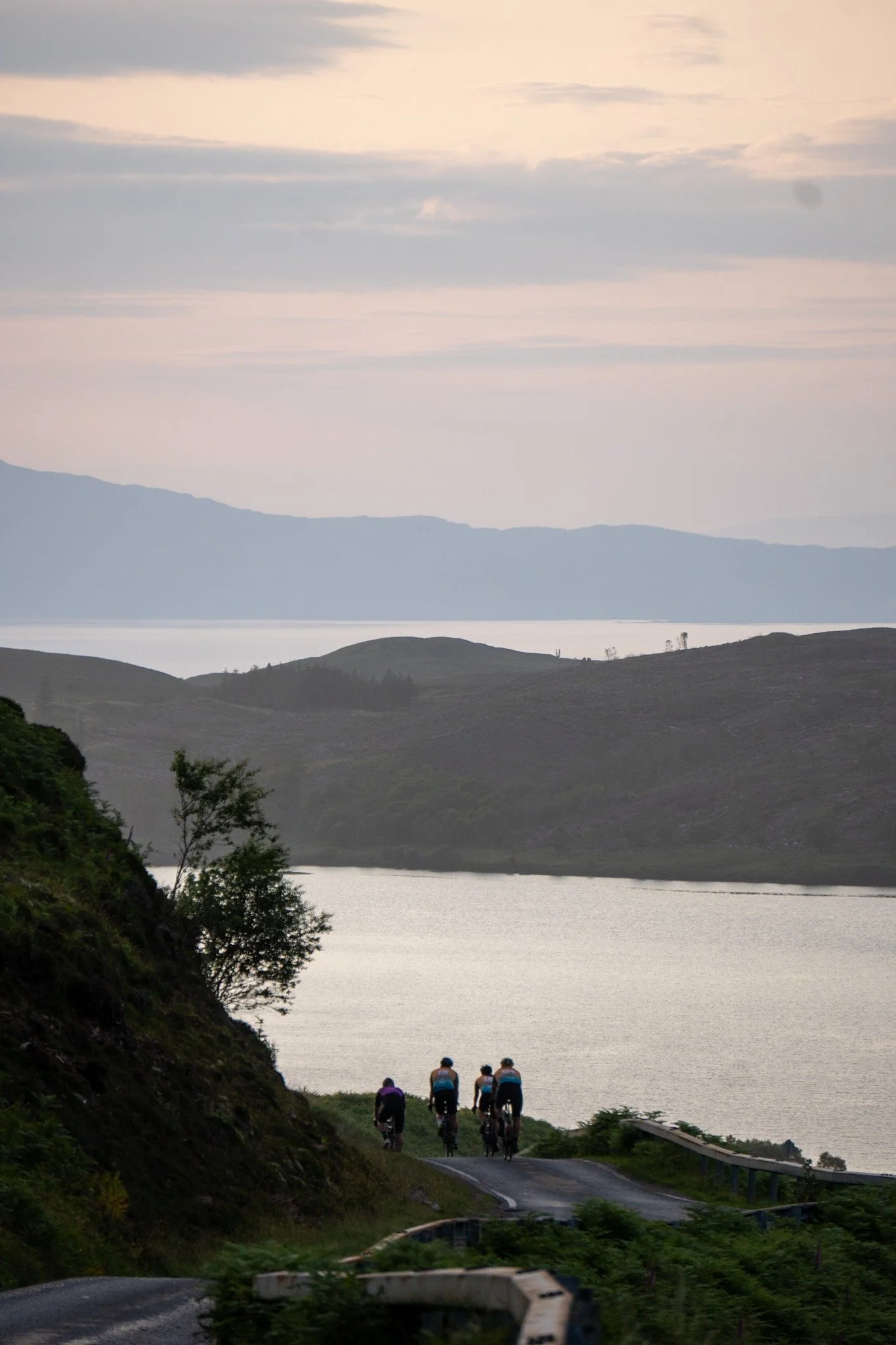 Four cyclists riding on a winding road along a hilly landscape with a body of water and mountains in the background during dusk.