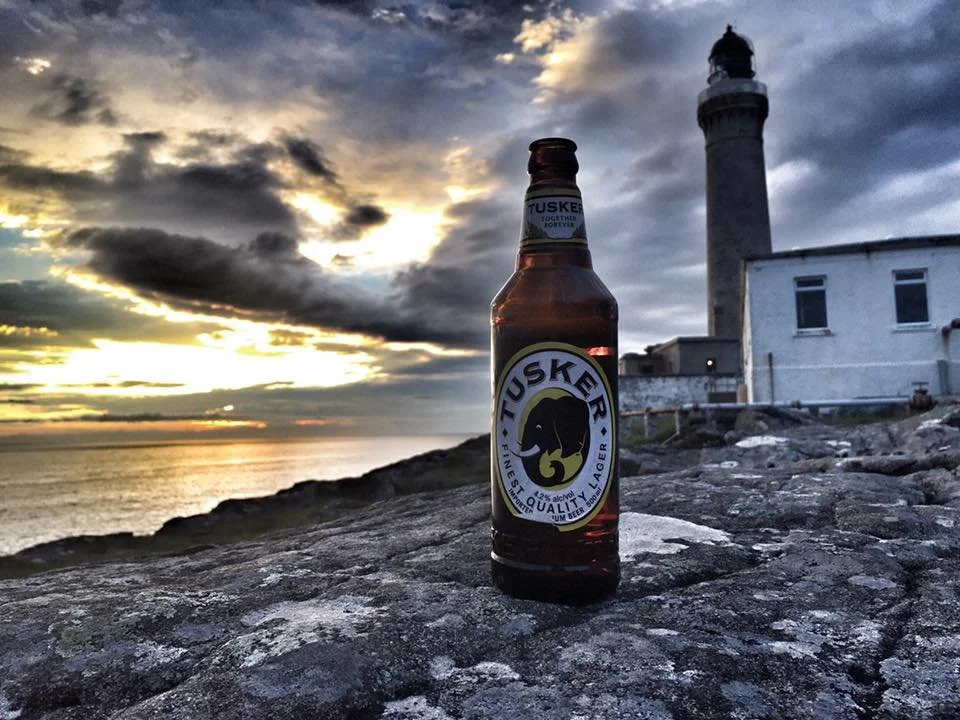 A bottle of Tusker beer placed on a rocky surface near a lighthouse, with a sunset and cloudy sky in the background.