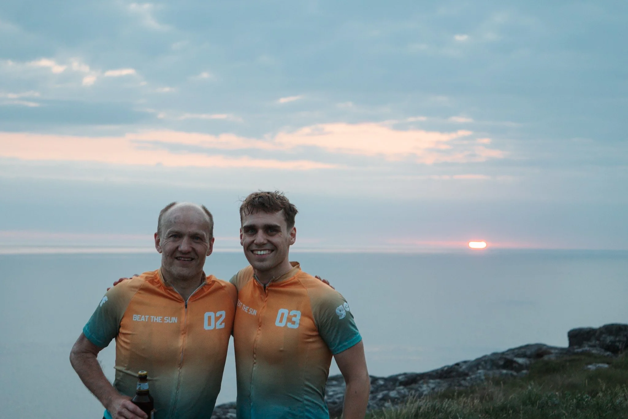 Two men in matching orange and blue cycling jerseys smiling outdoors with a sunset over water and rocky shoreline in the background.