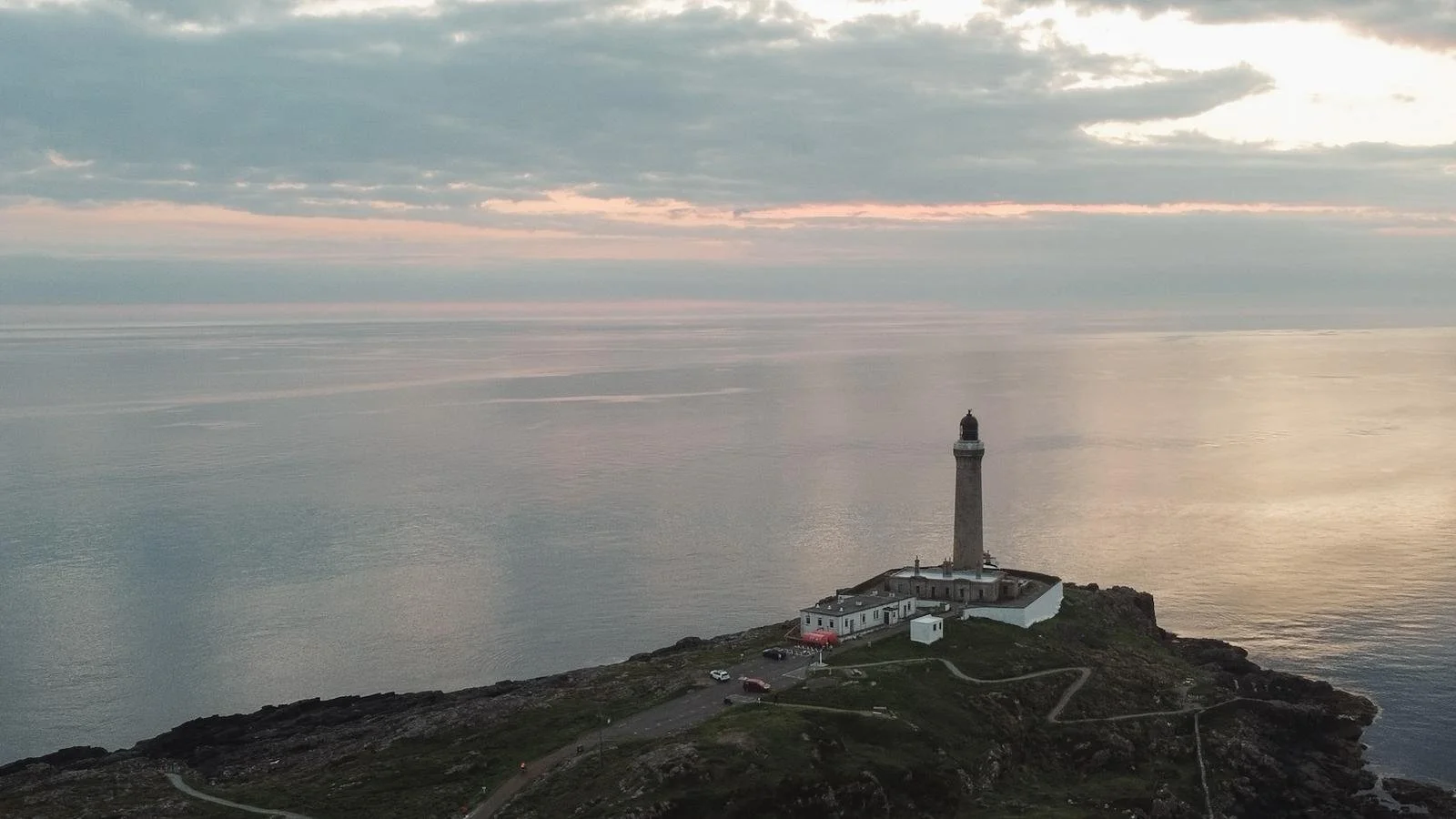A lighthouse on a rocky headland overlooking the ocean at sunset, with a cloudy sky and calm water.