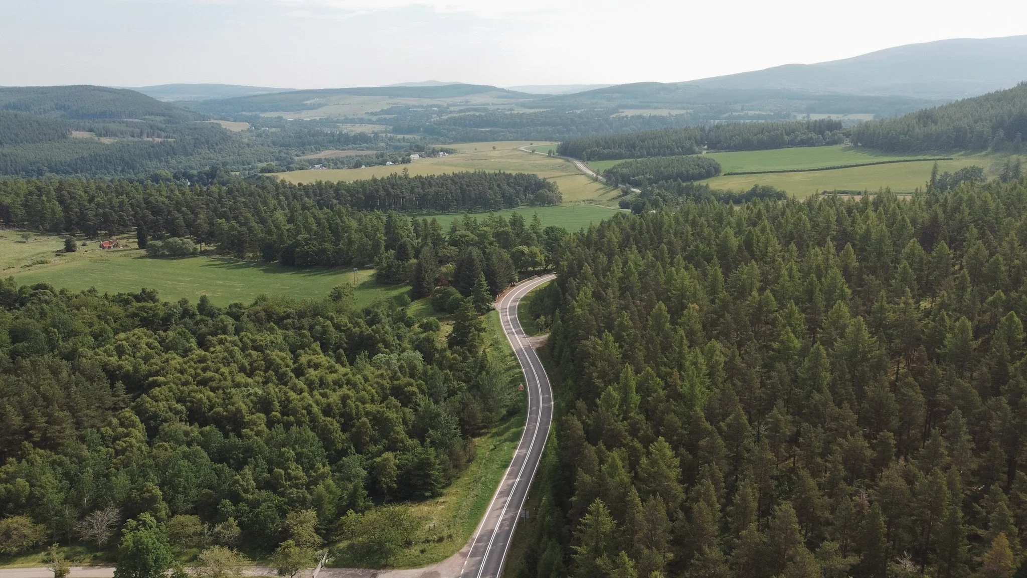 A winding road running through a lush green forest in a hilly landscape, with rolling hills and fields in the distance under a partly cloudy sky.