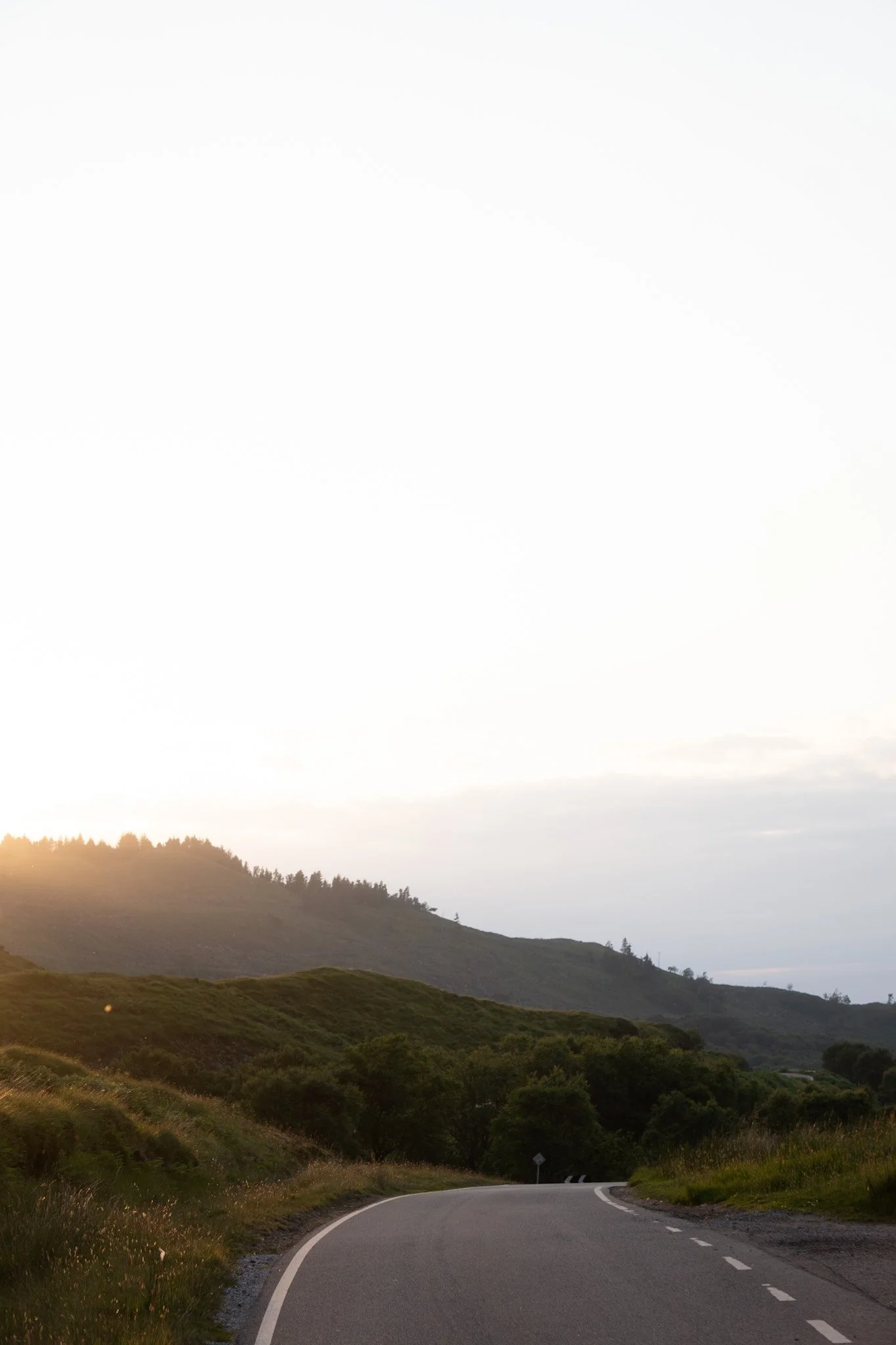 A winding country road surrounded by green hills with trees, under a partly cloudy sky during sunset.