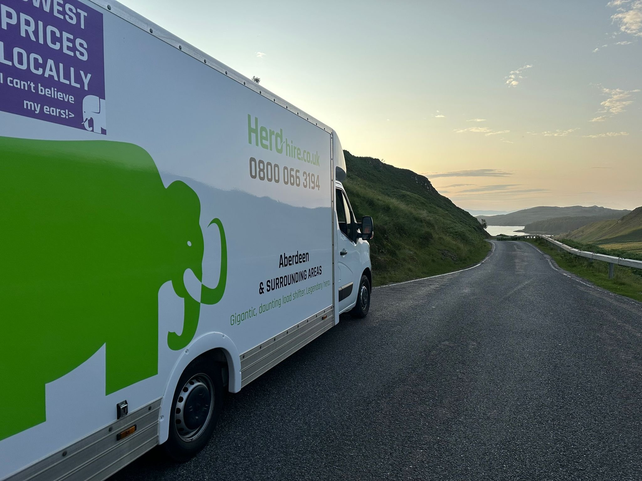 A white delivery truck with green and black text parked on a winding rural road at sunset, with hills and a body of water in the distance.