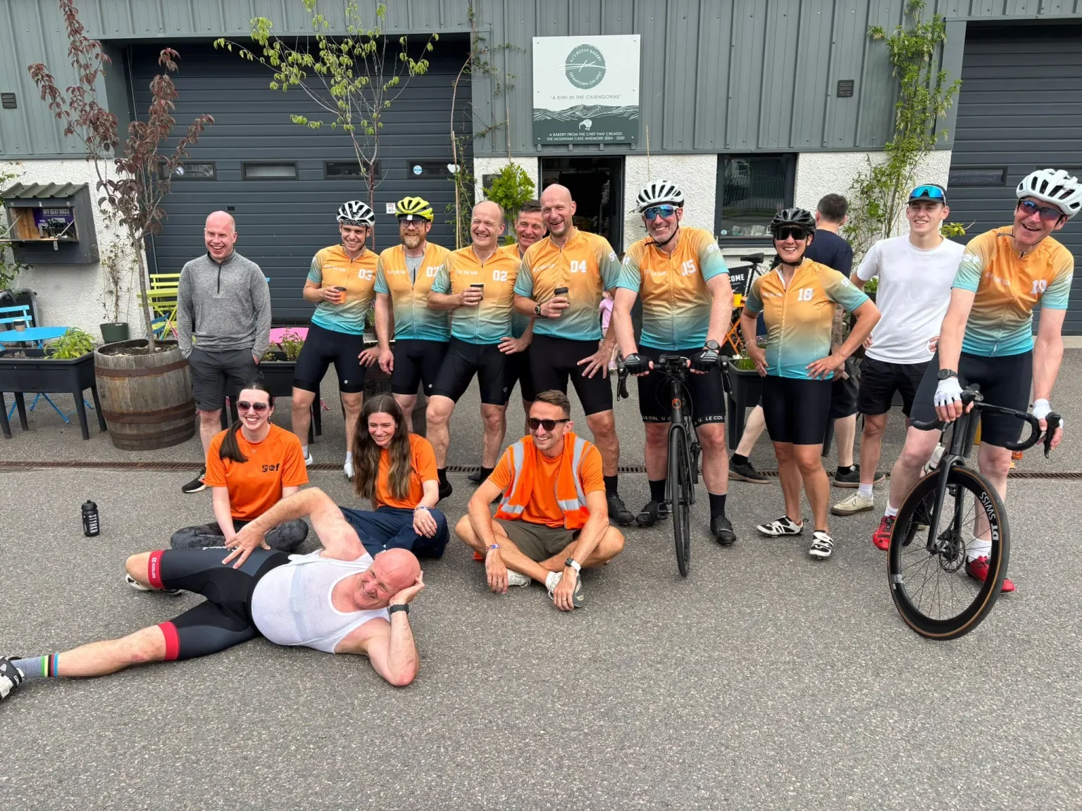 Group of cyclists posing outdoors in front of a gray building with plants, some are sitting on the ground, others standing, some wearing helmets and cycling gear, with a few holding drinks, smiling and enjoying a social moment.