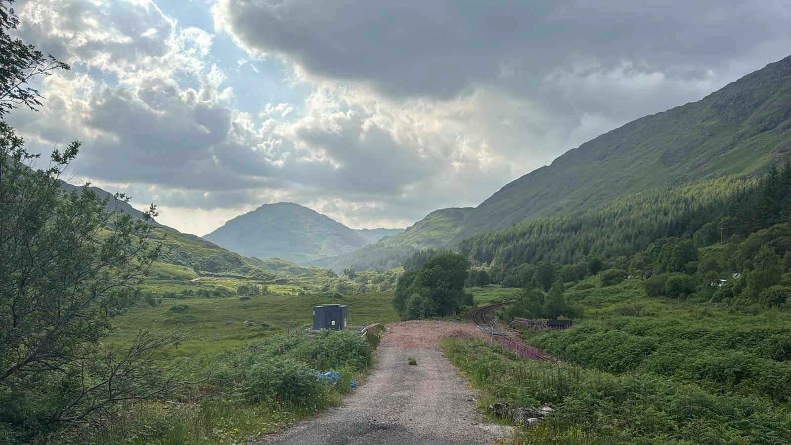 A scenic valley surrounded by green mountains, with a gravel path running through it and a small blue building on the left, under a cloudy sky.