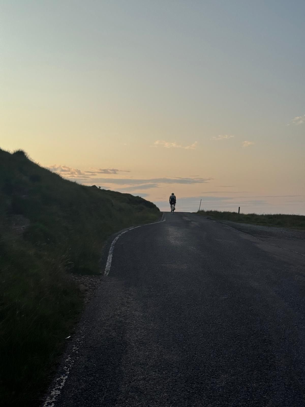 Person riding a bike on a winding road near the coast during sunset, with hills on the left and the ocean on the right.