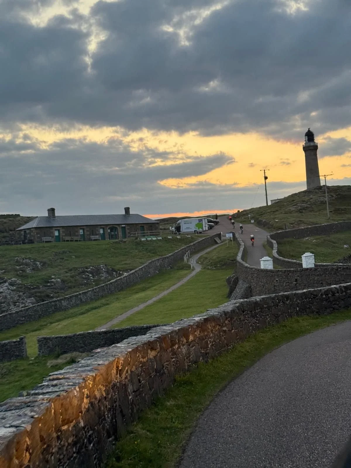 Scenic view of a lighthouse on a hill at sunset with winding paths, stone walls, a house, and motorcycles on the road.