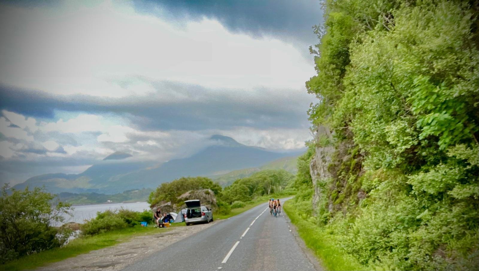 A scenic roadside view with a lake on the left, surrounded by green trees and mountains under a cloudy sky. A group of cyclists is riding on the road, and there are campers and tents near the water.