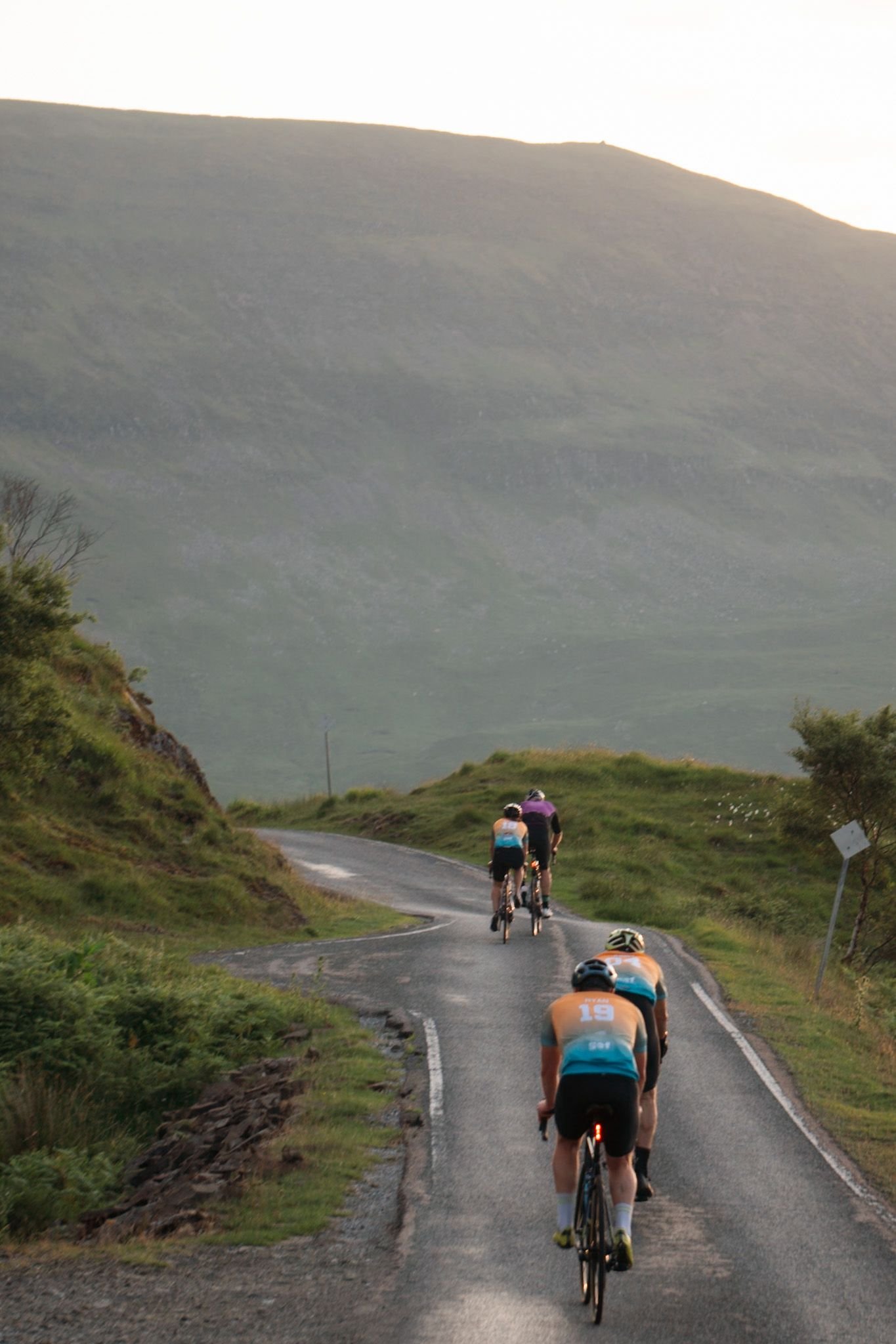 Four cyclists riding on a winding road through a green, hilly landscape during daytime.