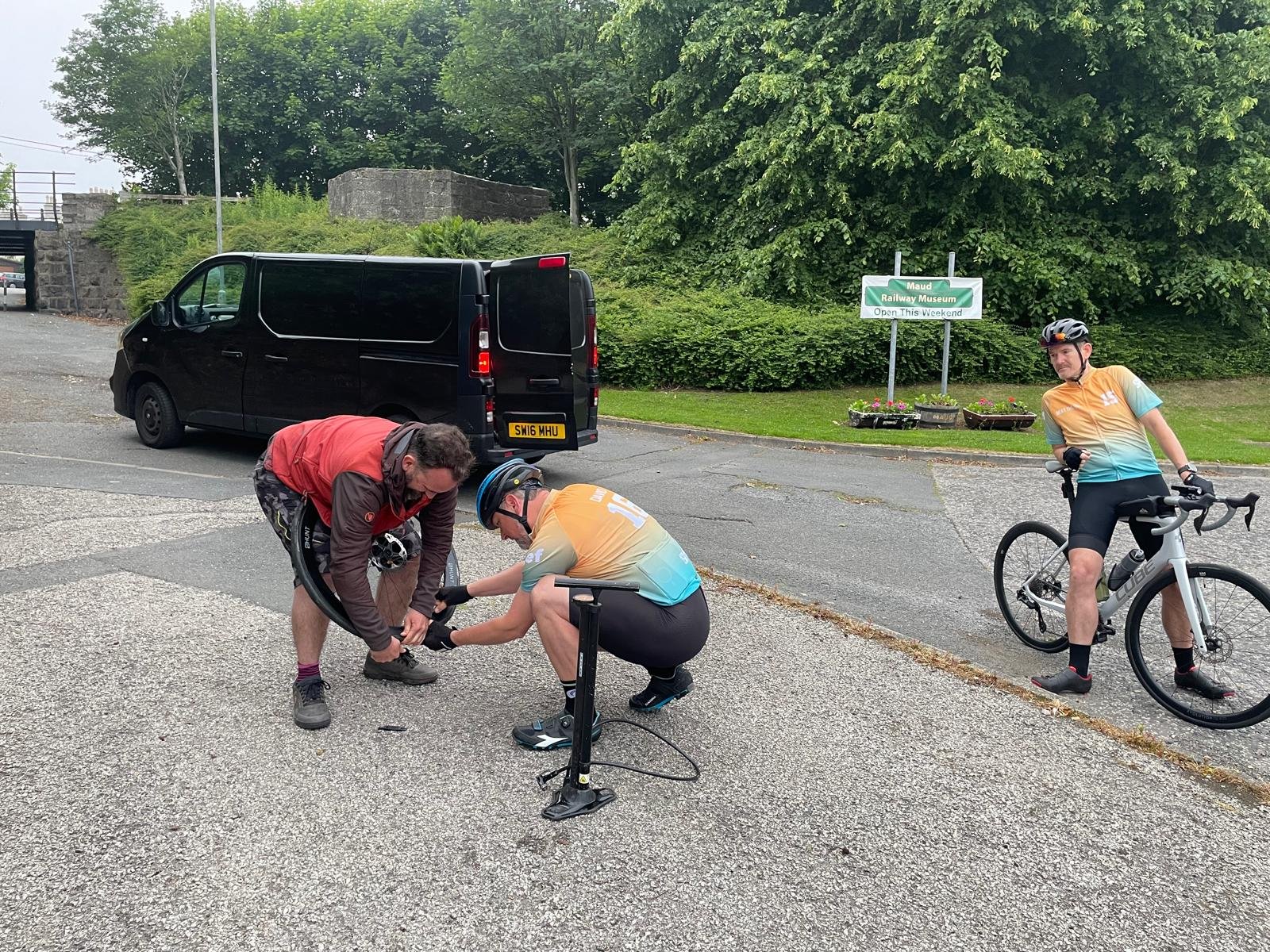 Two cyclists are assisting a third cyclist who is fixing a flat tire on his bicycle. One cyclist is squatting, the other is bending over holding the bike, and the third is standing nearby holding his bike and watching.