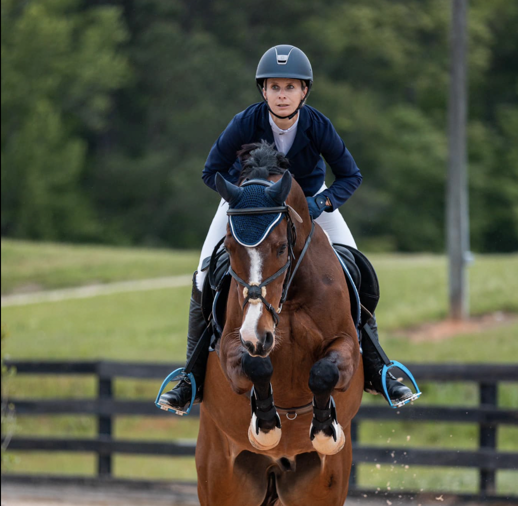 A woman wearing a helmet and riding gear, riding a brown horse with a blue ear bonnet, jumping over an obstacle.