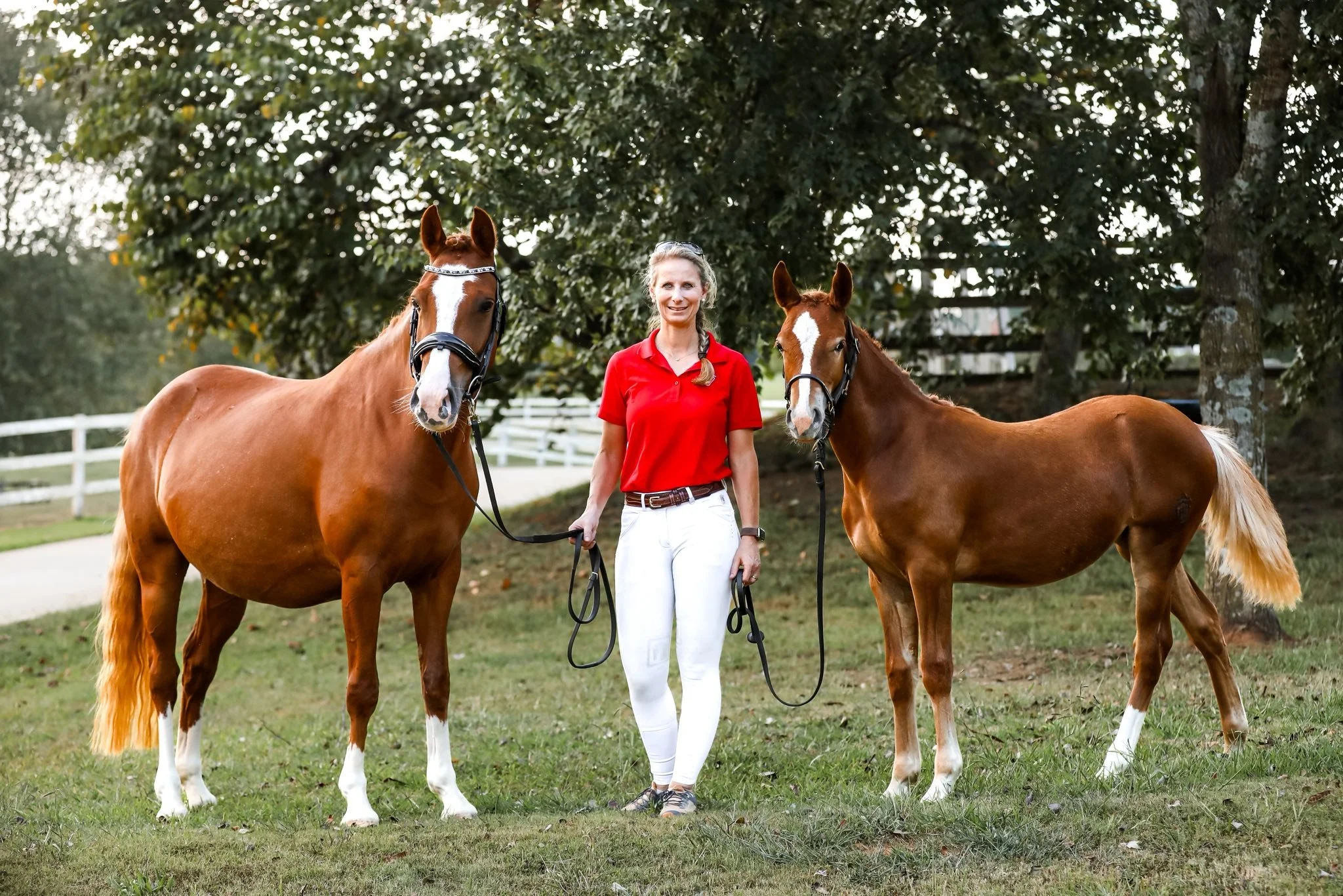 A woman standing outdoors holding two brown horses by their reins, with trees and a white wooden fence in the background.
