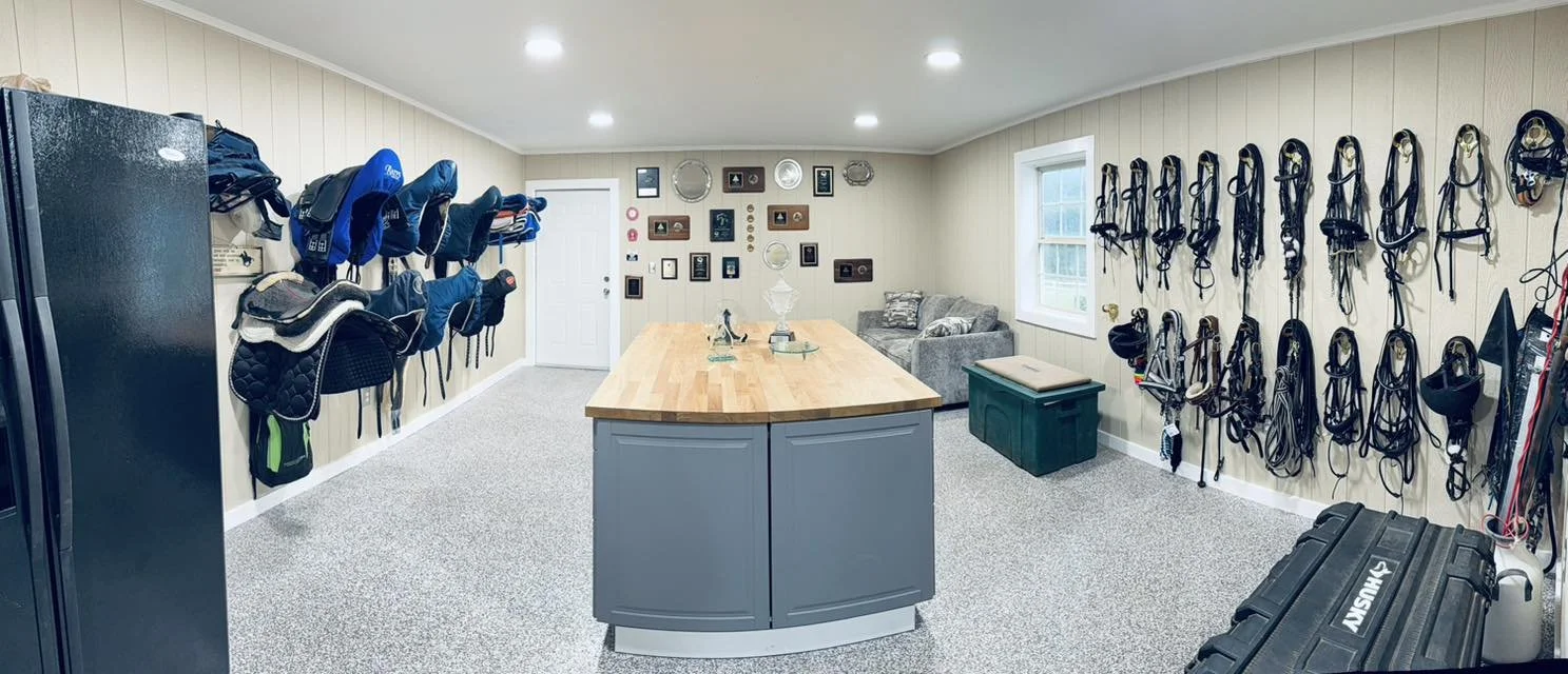 A spacious tack room with beige walls, a wooden table at the center, and equipment including horse bridles and saddles hanging on the walls.