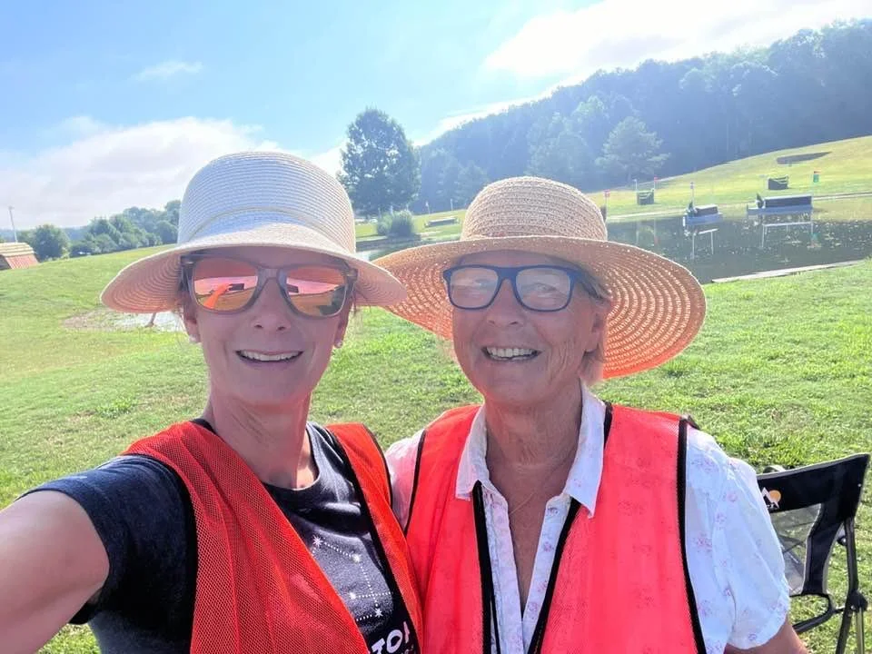 Two women wearing sun hats, sunglasses, and safety vests taking a selfie while volunteering at a horse show.