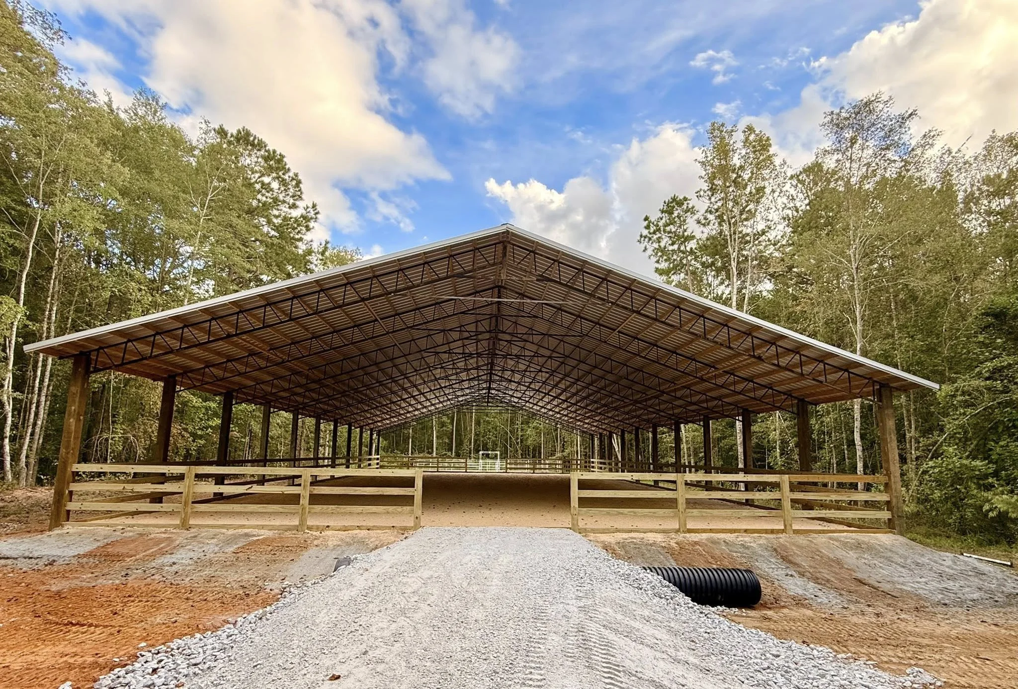 Construction site showing a large covered horse riding arena with metal roof supported by wooden beams, surrounded by trees and a gravel pathway in the foreground.