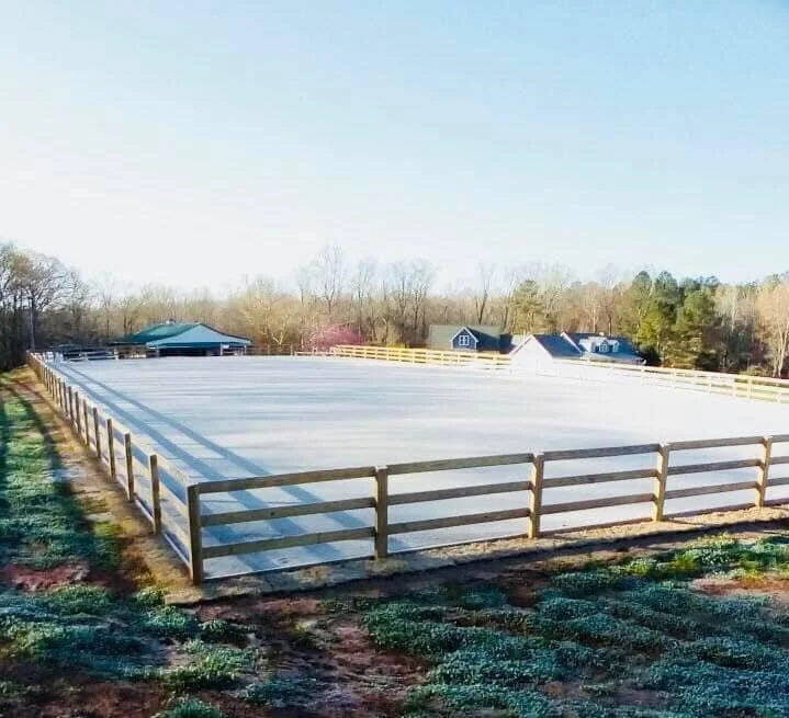 An outdoor horse riding arena surrounded by a wooden fence, with trees, houses, and a blue sky in the background.