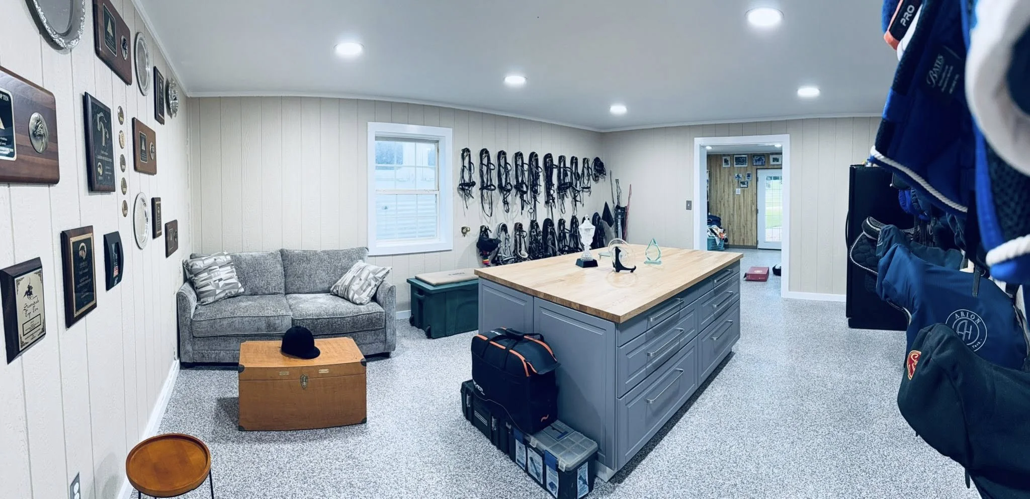 Tack room with awards on wall, gray sofa with pillows, wooden island, large window, tool and gear storage, central island with a wooden top, black bag under the island, and sports equipment on the right wall.